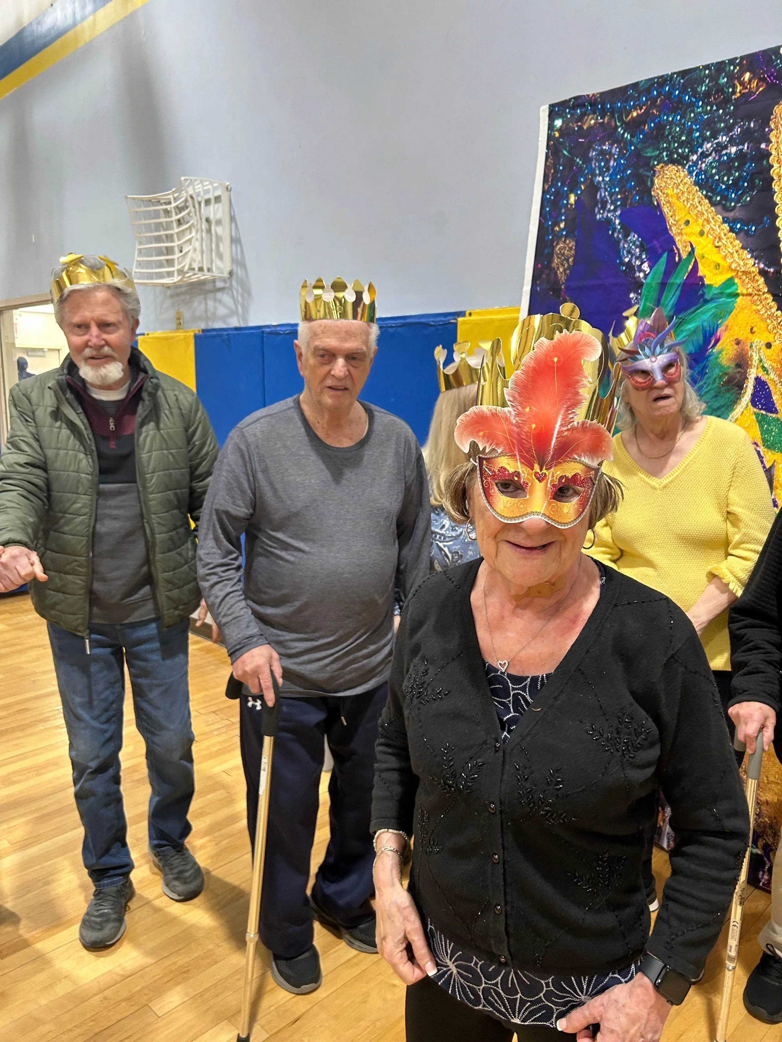 A group of five elderly people celebrating Mardi Gras, some wearing colorful masks and gold crowns, in a room with wooden floors and a large colorful artwork in the background.
