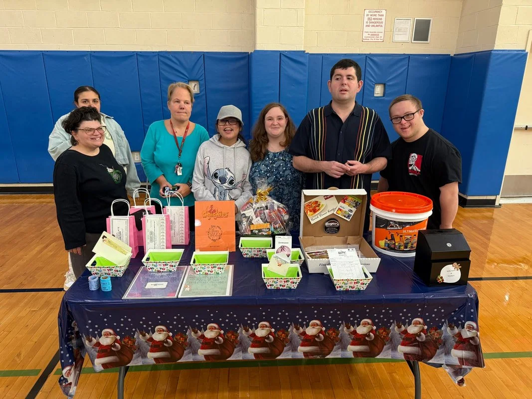 Group of people standing behind a table decorated with a Christmas-themed tablecloth, displaying gift bags, boxes, and holiday items, in a gymnasium.