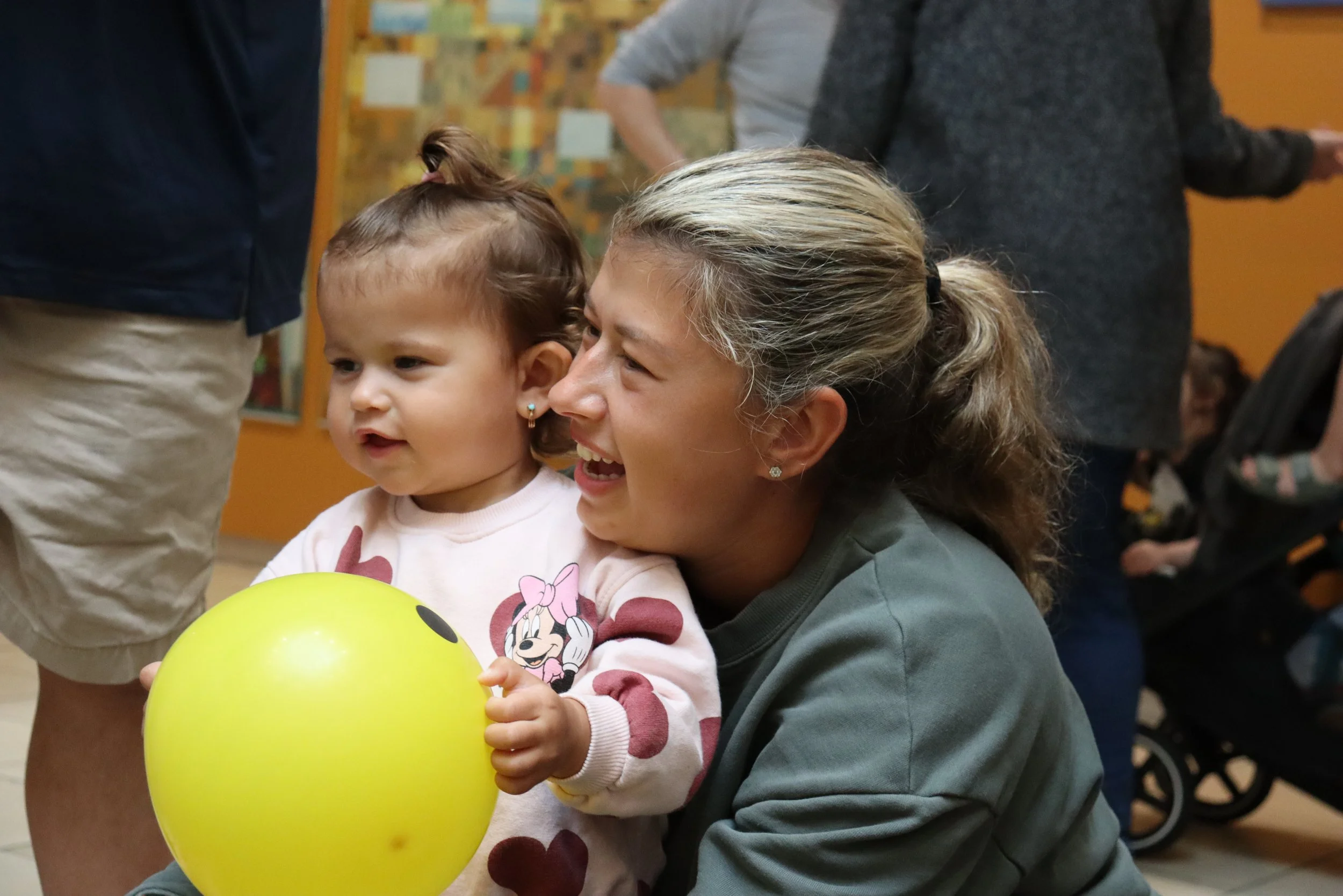 A young girl holding a yellow balloon with a smiley face, smiling and looking at a woman who is crouching and also smiling. The girl wears a pink Minnie Mouse sweatshirt, and the woman has blonde hair tied back and wears a grey hoodie. There are other people in the background, indicating a social setting.