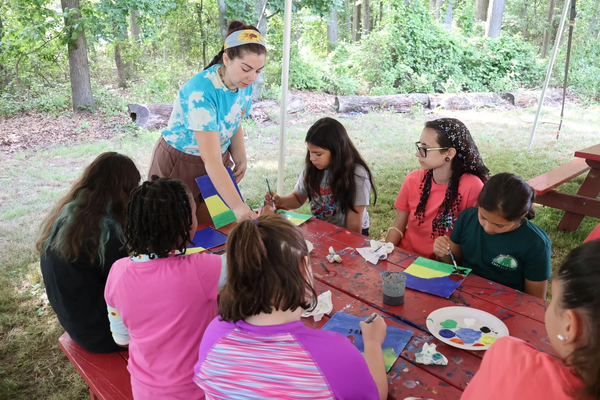 A group of diverse children sitting around a red picnic table outdoors, painting on paper with an adult woman supervising. The setting is a forested area with trees and greenery.