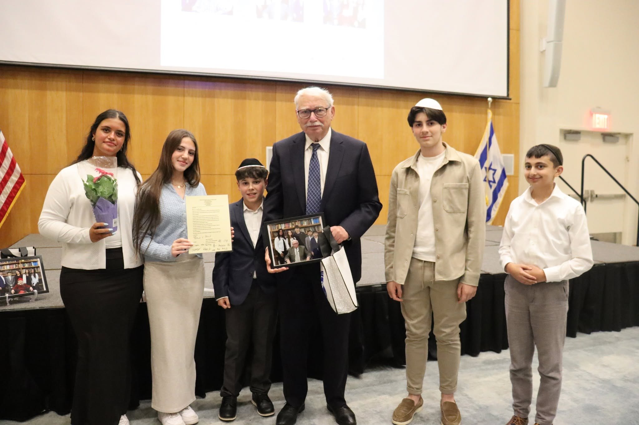 Group of six people, including five children and one older man, standing indoors in front of a wooden wall with a projector screen showing small images. The children are holding certificates, flowers, and a framed photo, and they are dressed in formal and semi-formal attire. An American flag and a blue and white flag are visible in the background.