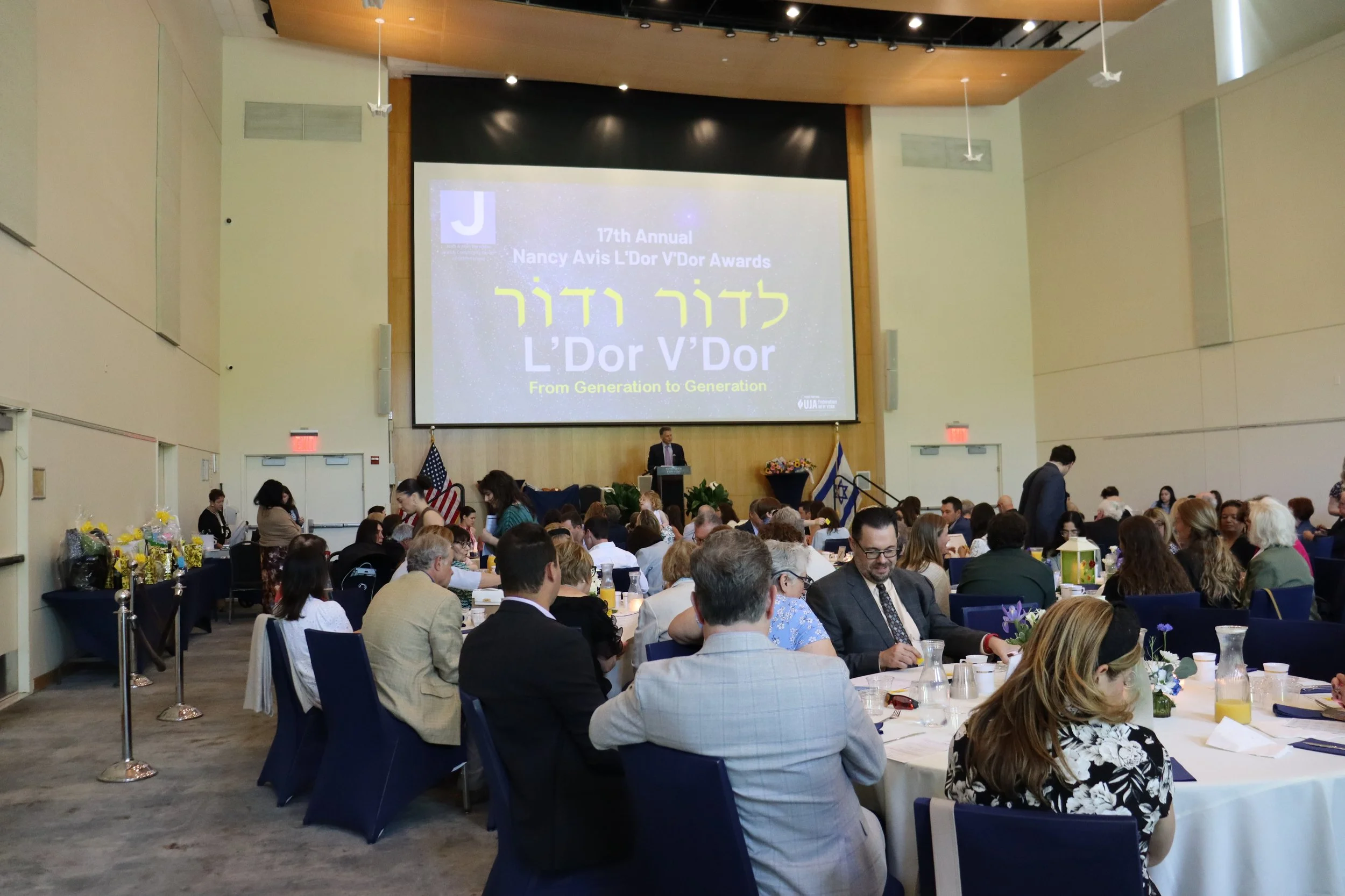 A large conference hall filled with people sitting at tables during an awards ceremony. A stage at the front holds a speaker at a podium, with a large screen displaying the event details: 77th Annual Nancy Avis L'Dor V'Dor Awards from Generation to Generation. The hall is decorated with flowers and flags, including the American and Israeli flags.