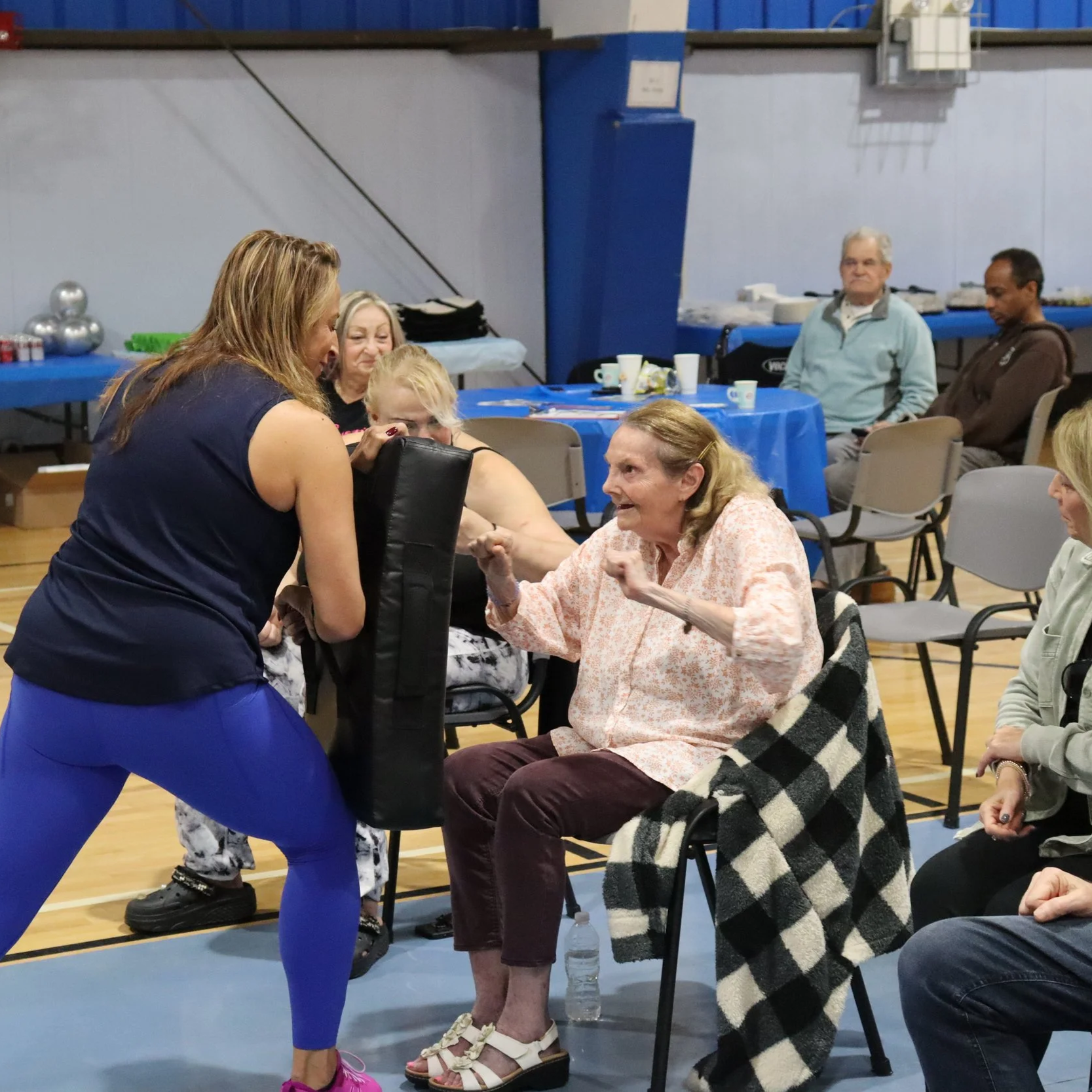 A group of people gathered in a room with tables and chairs, engaging in an arm wrestling match between an elderly woman and a younger woman, with others watching nearby.