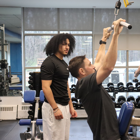 A person demonstrating a strength training exercise on cable machine while a trainer observes in a gym.
