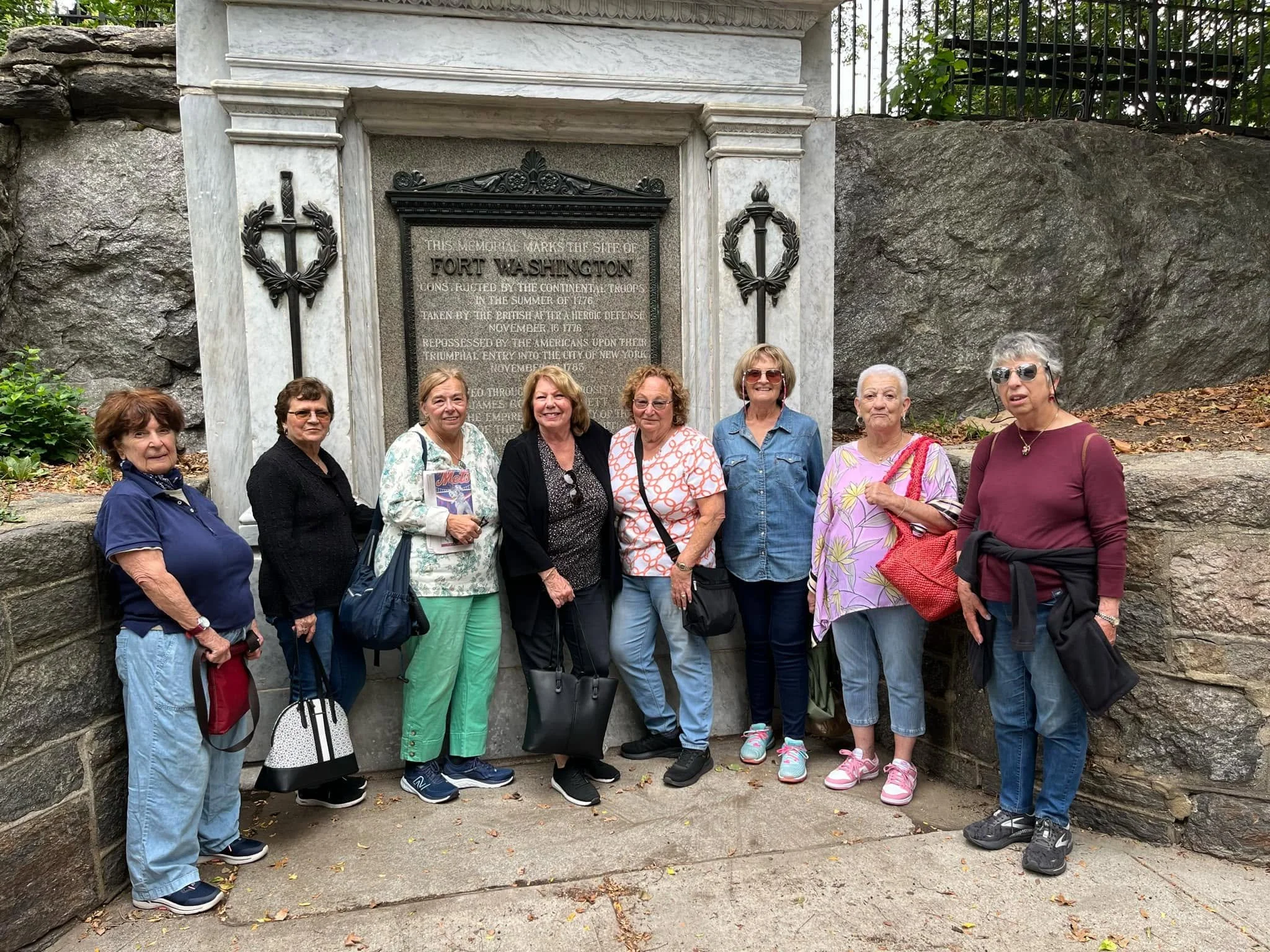 Group of women standing in front of a memorial marker for Fort Washington, which is embedded in a rock wall, with greenery and black fence above.