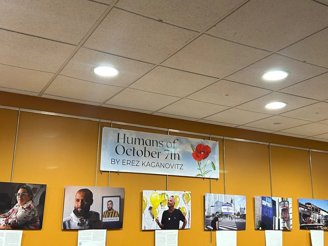 Exhibition display titled 'Humans of October 7th' by Erez KaganoVitz, featuring portraits of diverse people with descriptive text below, set against a yellow wall.