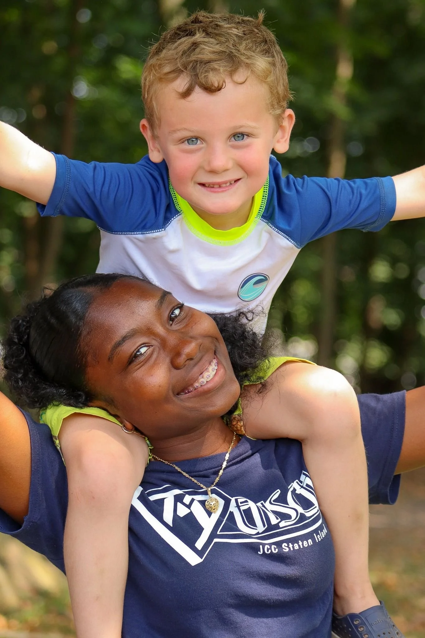 A young boy with curly blonde hair and blue eyes is smiling, balancing on a woman's shoulders with her smiling face partially visible, outdoors with green trees in the background.