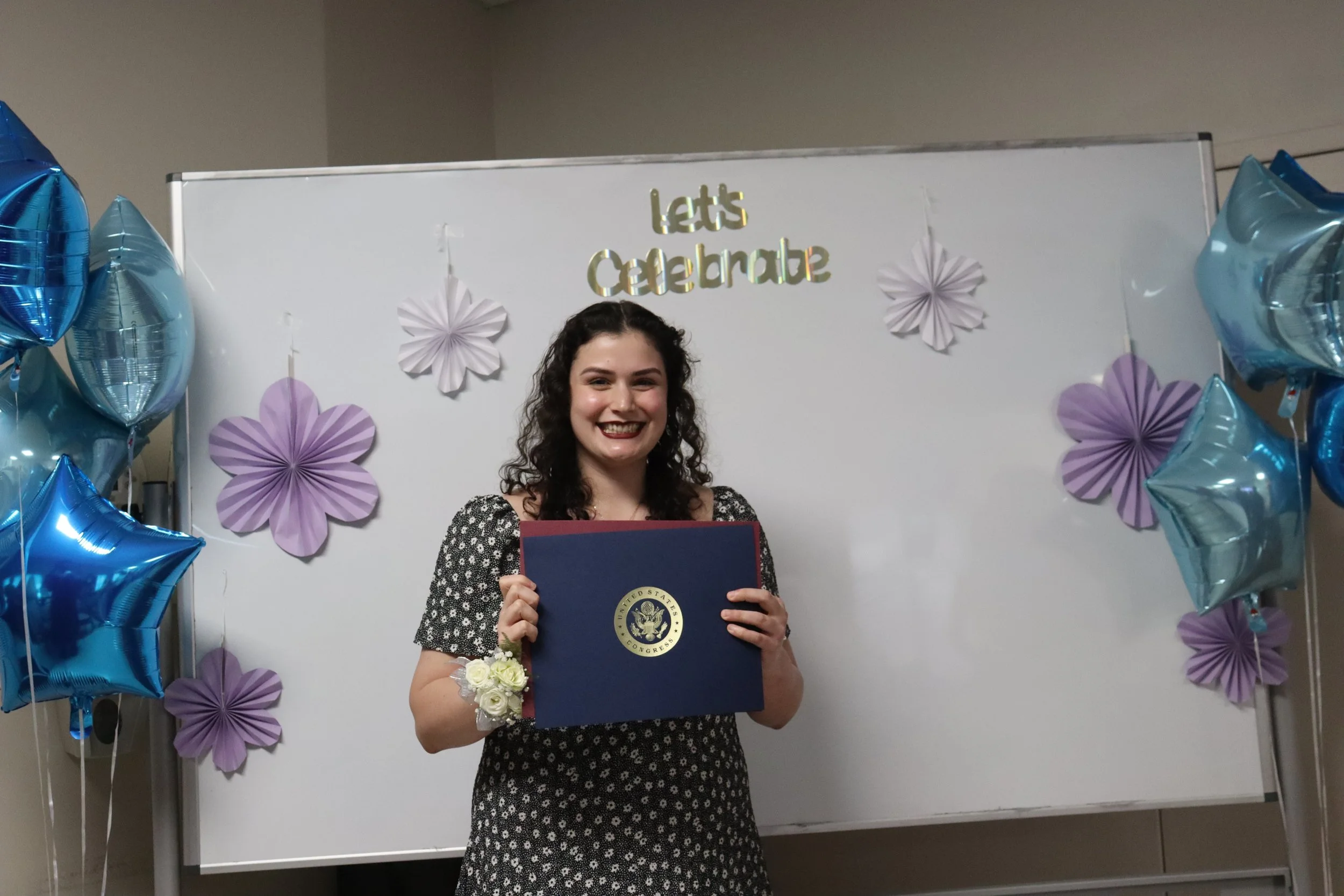 A young woman with curly hair holding a diploma on graduation day, surrounded by purple and blue balloons and decorations on a white backdrop with the words 'Let's Celebrate' in gold lettering.