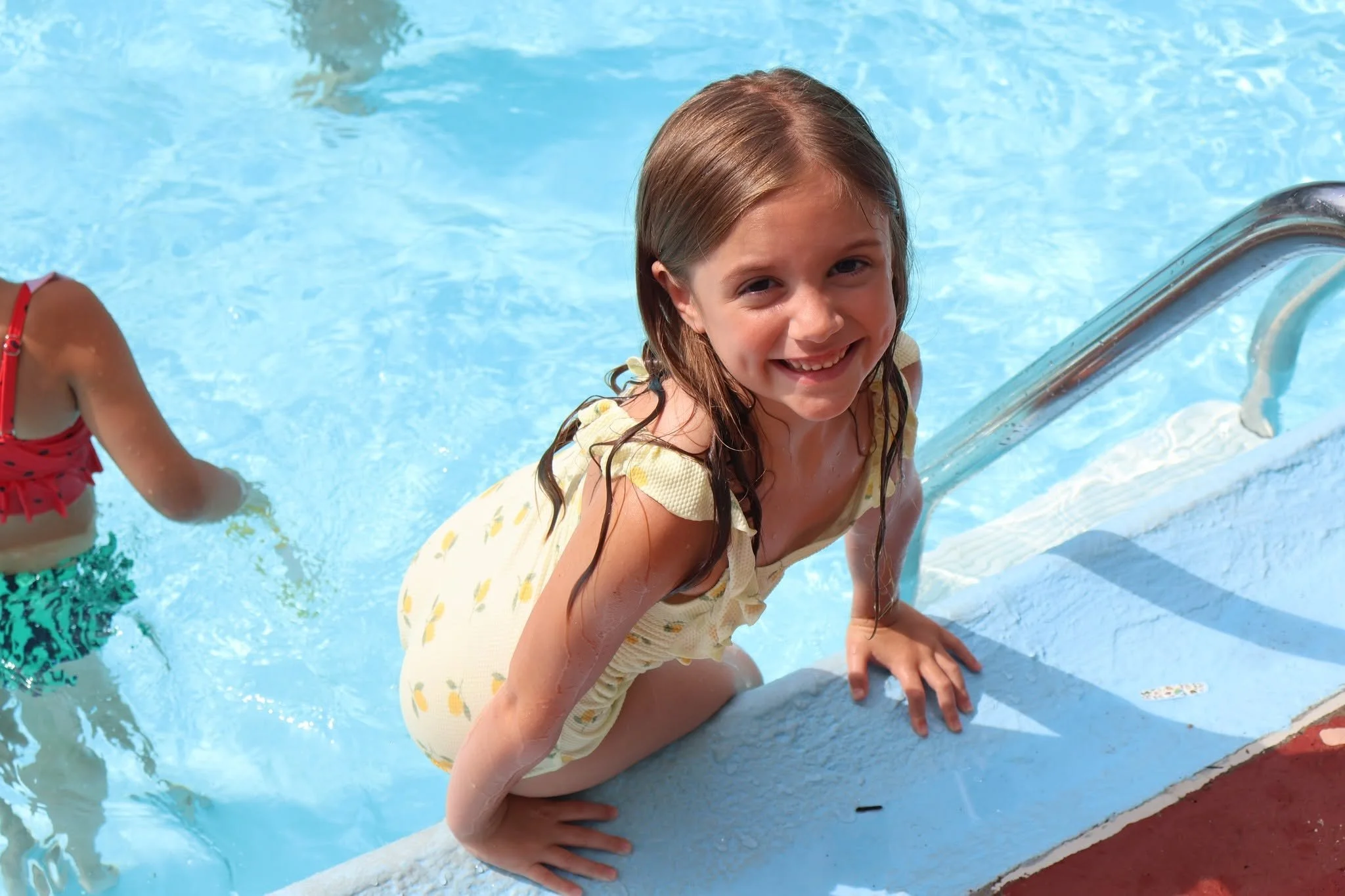 Smiling girl in a yellow dress with lemon print squatting by the edge of a swimming pool, holding onto the pool deck with her hands.