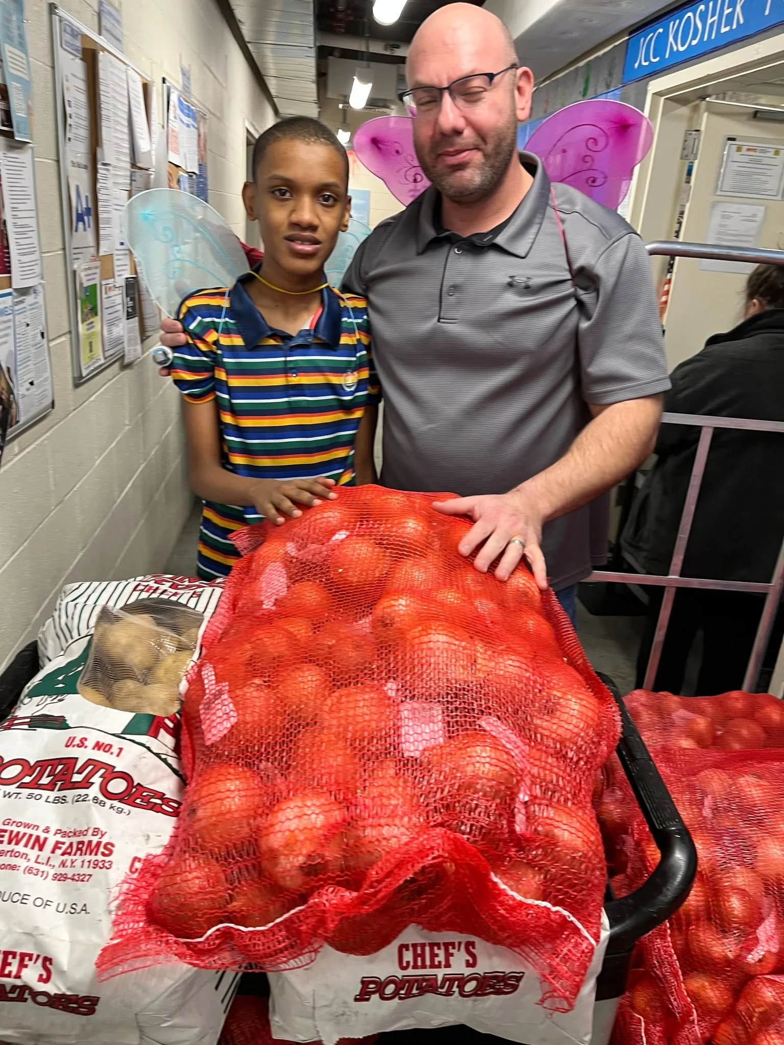 A man and a young boy with fairy wings posing behind a large red mesh bag of onions, in a store with papers on the wall and people in the background.