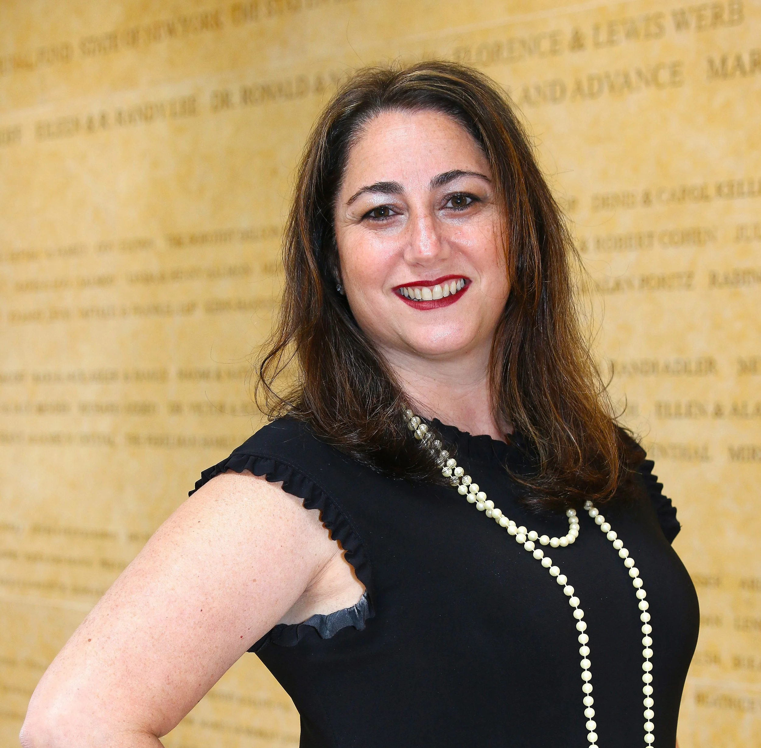 A woman with brown hair and fair skin smiling at the camera, wearing a black top with ruffled sleeves and a long string of pearls, standing in front of a beige background with text.