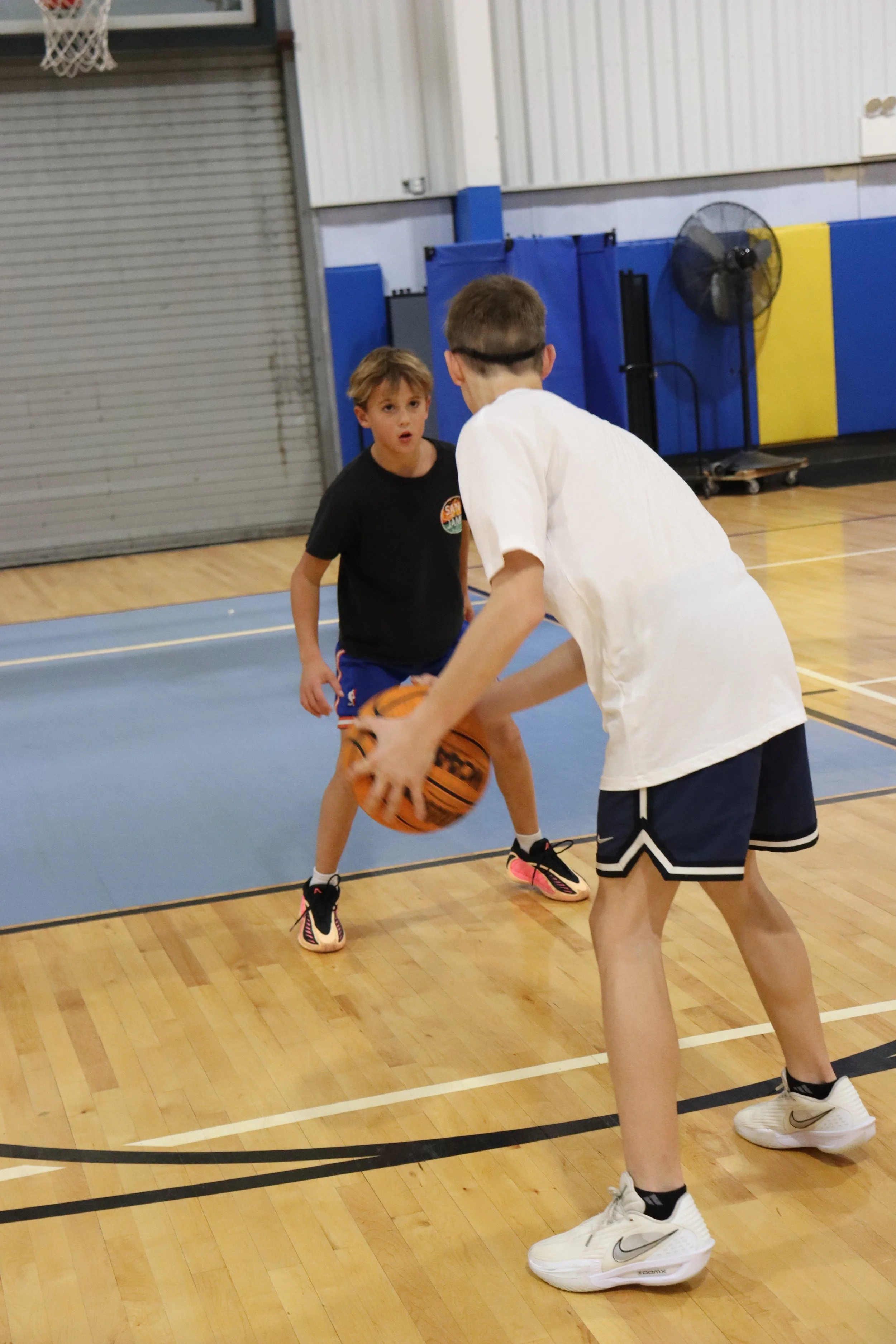 Two boys playing basketball in an indoor gym, one wearing a white shirt and shorts, the other in a black shirt and blue shorts, with one boy holding a basketball and the other defending.