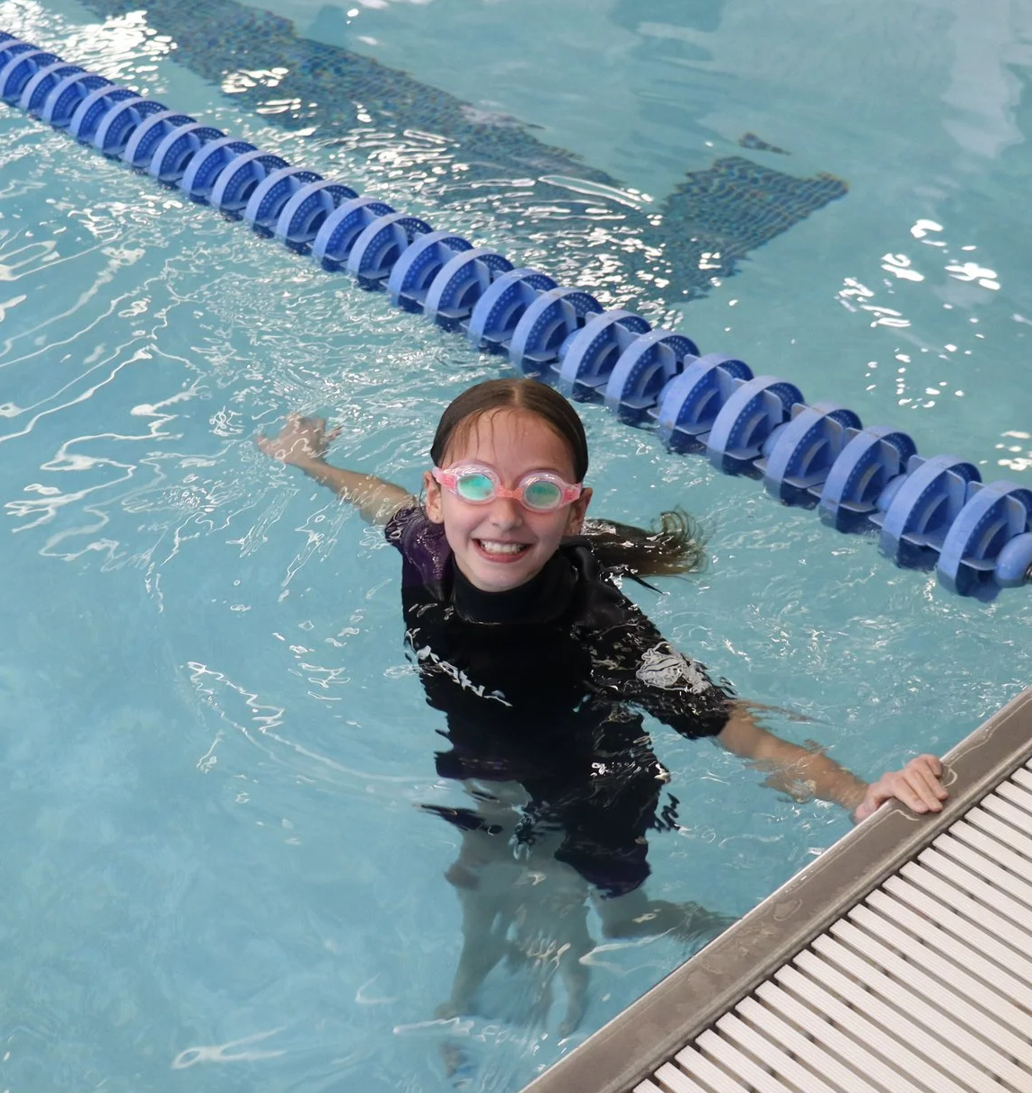 A young girl wearing goggles and a black swimsuit smiling in a swimming pool, holding onto the pool edge with one hand and floating near the lane divider.