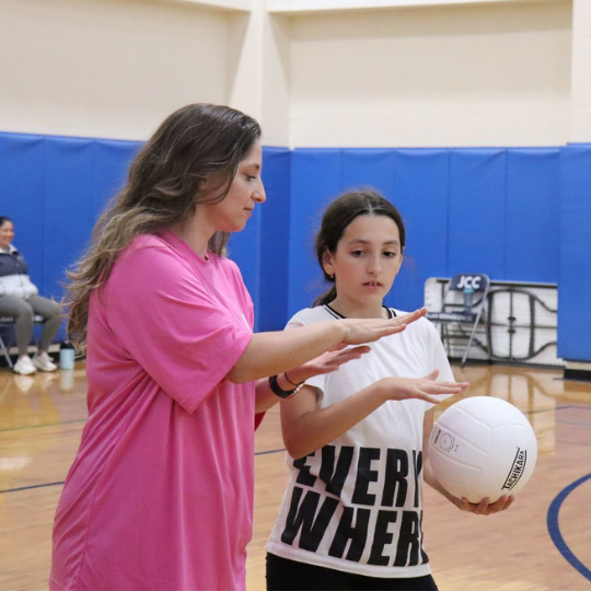 A woman in a pink shirt coaching a young girl in a white sports shirt with black lettering, holding a volleyball, in an indoor gymnasium with blue padded walls.