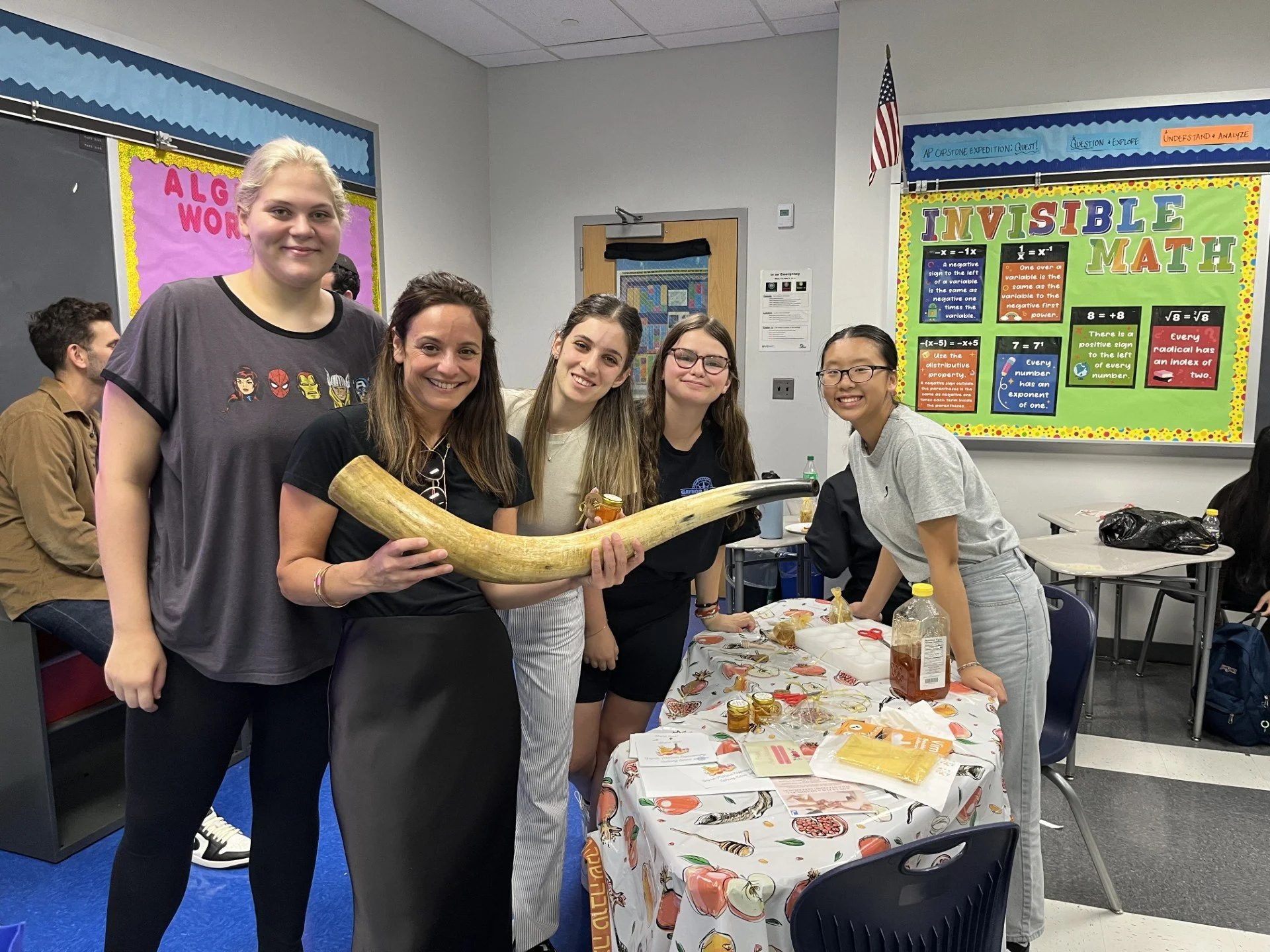 Group of five women standing around a table in a classroom, holding a large animal horn, with craft supplies on the table, colorful educational posters on the walls, and other students in the background.