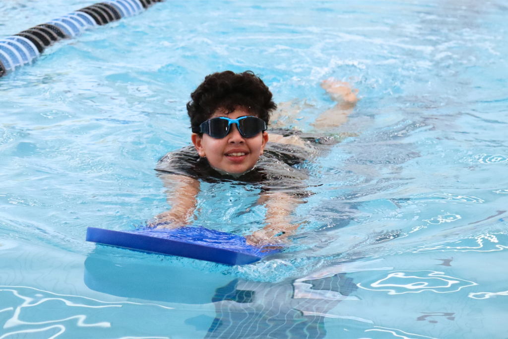 A young boy wearing goggles swimming in a pool and holding a blue kickboard.
