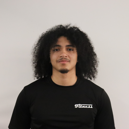 A young man with curly black hair and a thin mustache, wearing a black T-shirt with white text, standing in front of a plain light-colored wall.