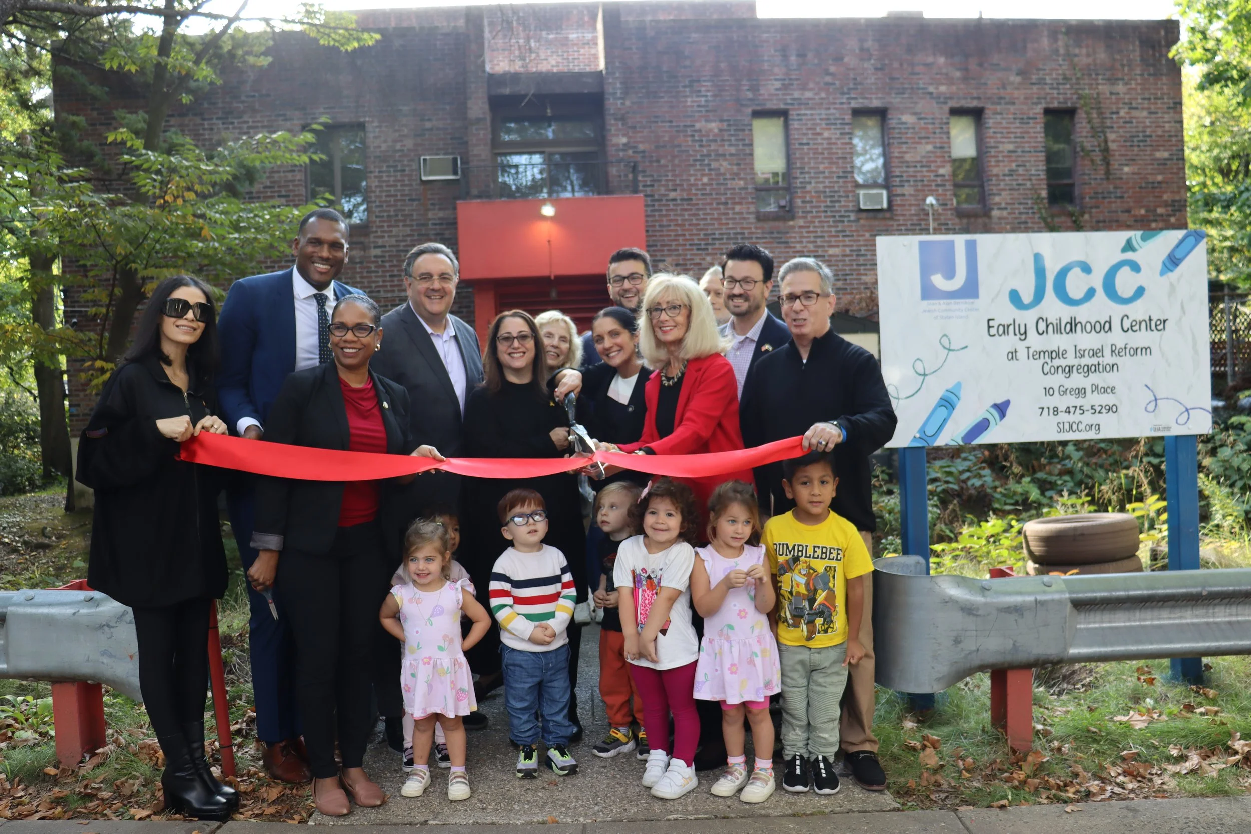 A diverse group of adults and children at a ribbon-cutting ceremony outside a brick building, with a sign indicating it is the Early Childhood Center at Temple Israel Reform Congregation. The group is smiling and gathered around a woman in a red blazer holding a pair of scissors, preparing to cut the red ribbon.