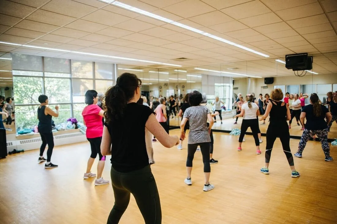 A group fitness class in a dance studio, with women following an instructor near the mirrors and large windows, practicing dance or aerobics.