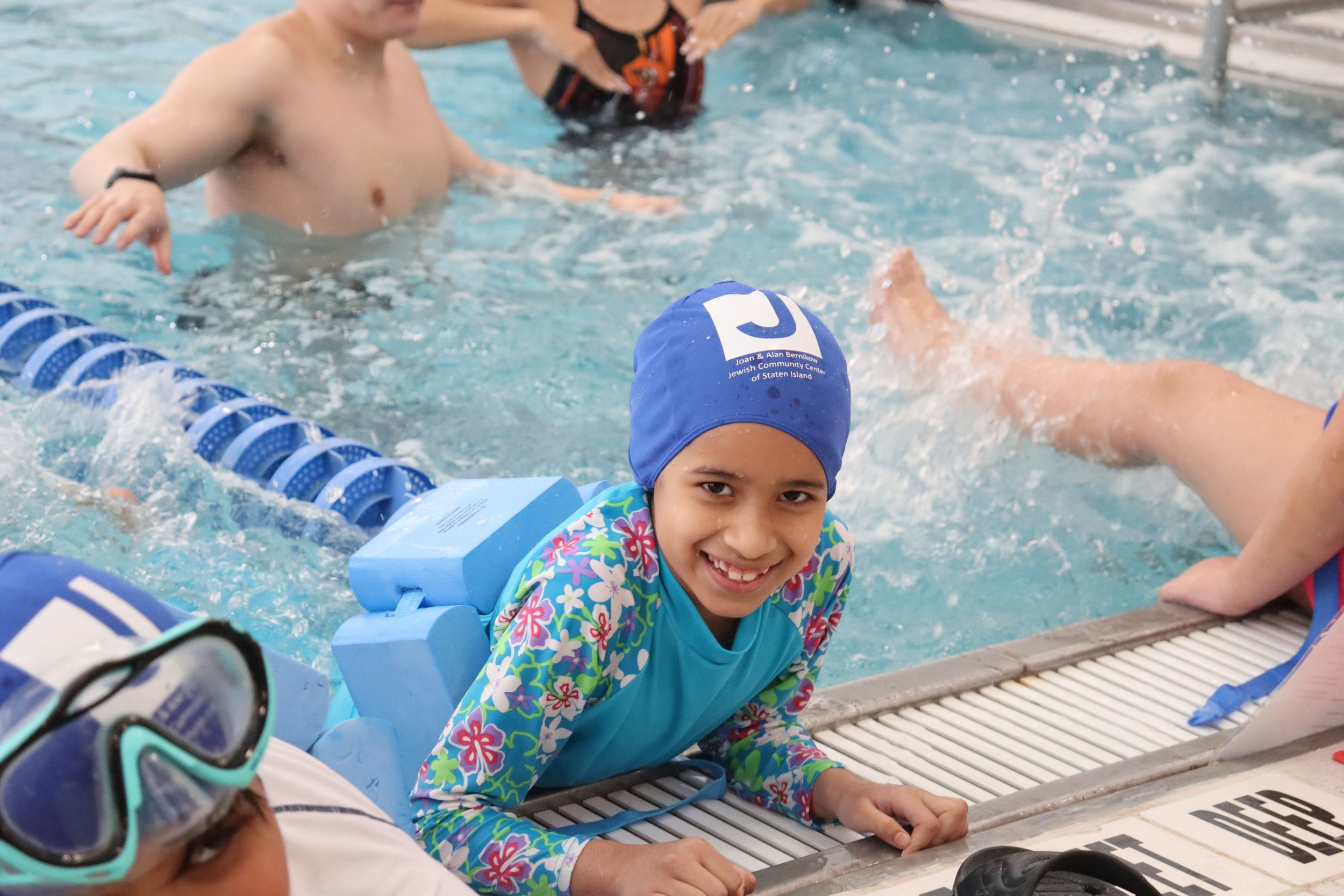 A young girl wearing a blue swim cap and floral swimsuit smiling while sitting at the edge of a swimming pool, with other children swimming in the background.