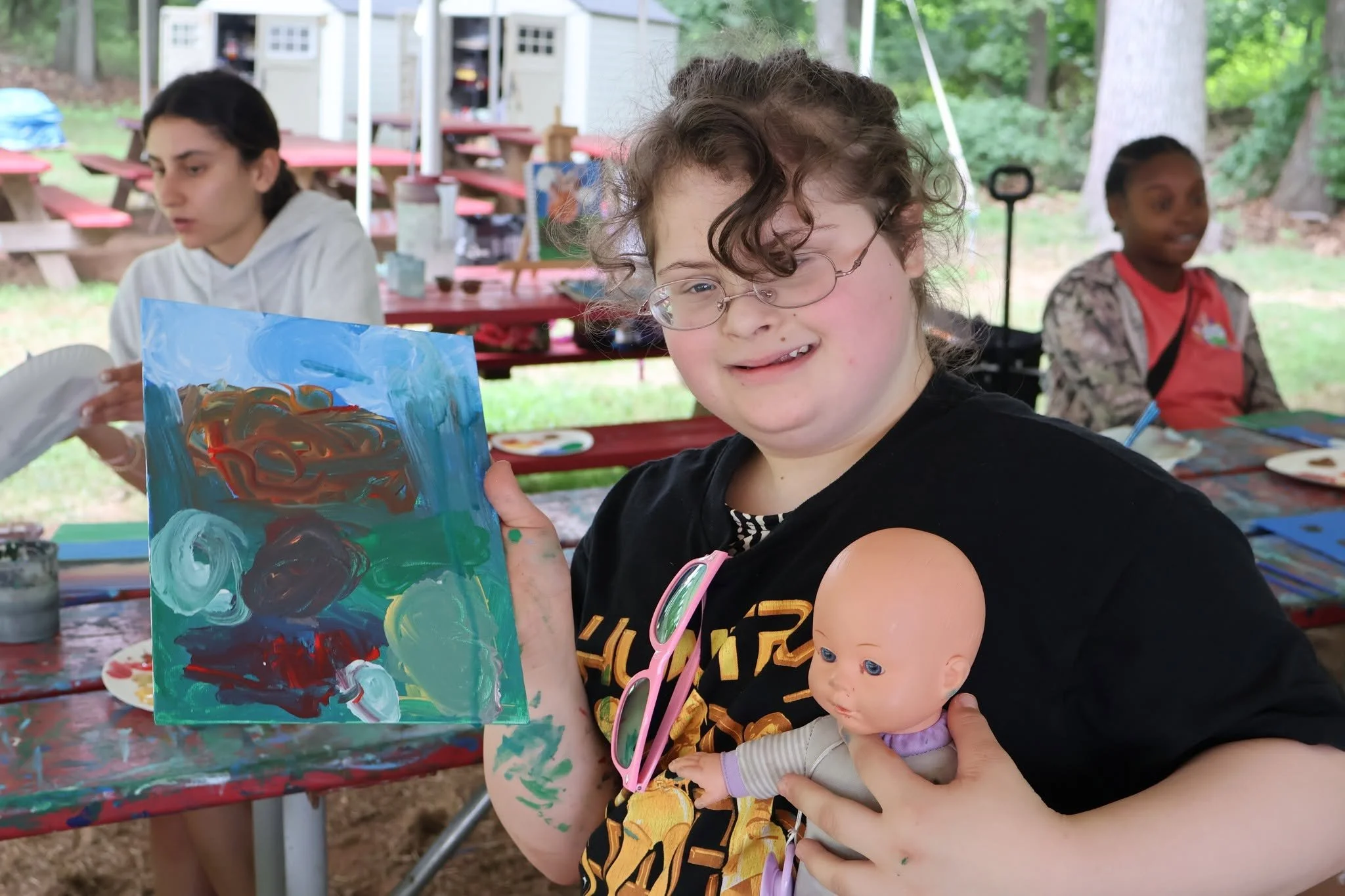 A young girl with curly hair and glasses holding a doll in one arm, showcasing her colorful abstract painting during an outdoor art activity.