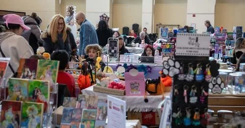 People shopping at a craft fair or market, browsing tables filled with handmade crafts, jewelry, and greeting cards.