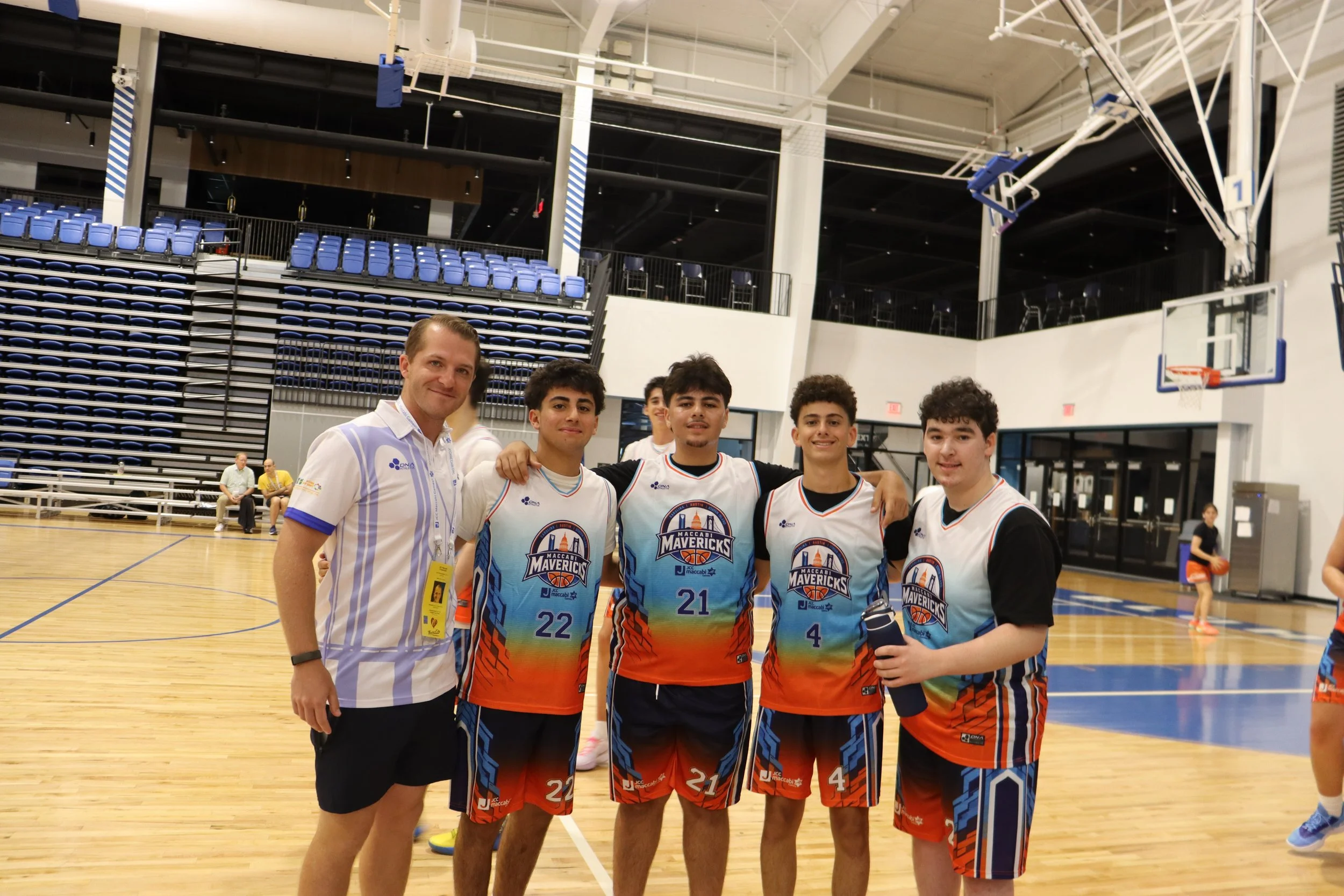 A group of five young basketball players and one adult posing for a photo on an indoor court. The players are wearing colorful, branded basketball uniforms with short sleeves and shorts. The adult is dressed in a striped shirt and black shorts. They are smiling, and some have their arms around each other. The background includes blue stadium seats, a basketball hoop, and a high ceiling in the sports complex.