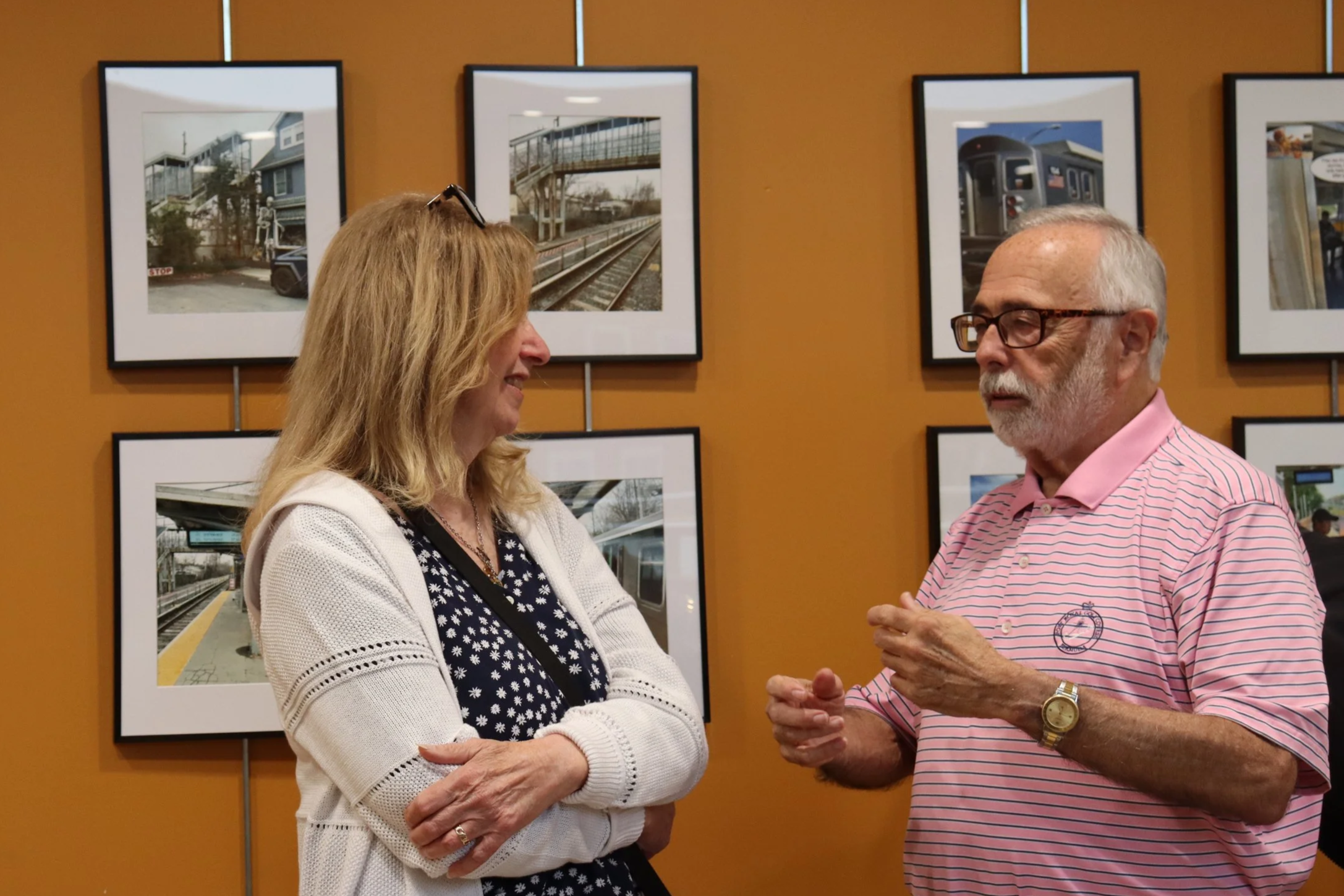 Two people, a woman with blonde hair and a man with gray hair and glasses, are talking to each other in an art gallery with framed photographs of train stations on an orange wall behind them.