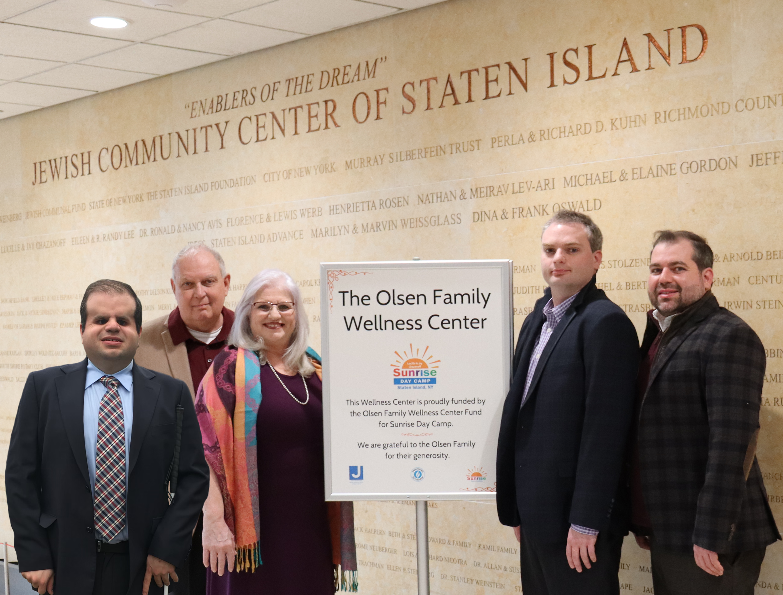 Group of five people standing in front of a large wall with text, about a wellness center and a donation from the Olsen Family during a daytime camp event on Staten Island, NY.