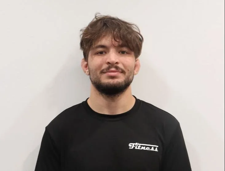 A young man with brown hair and a beard, wearing a black T-shirt with a white logo, standing against a white wall.