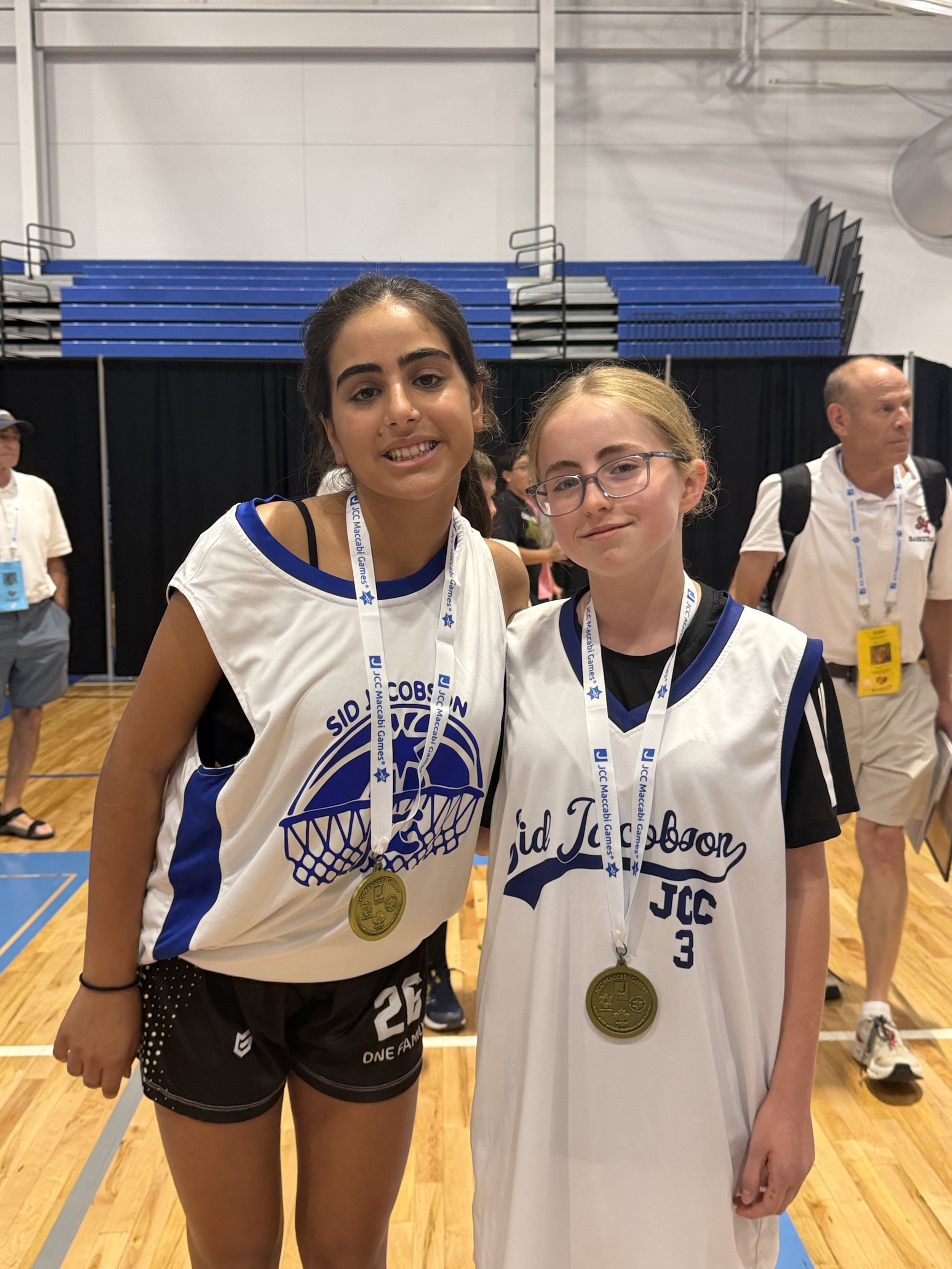 Two young female athletes with medals around their necks, wearing white and blue sports uniforms, standing on a basketball court after a competition with a packed gymnasium and spectators in the background.