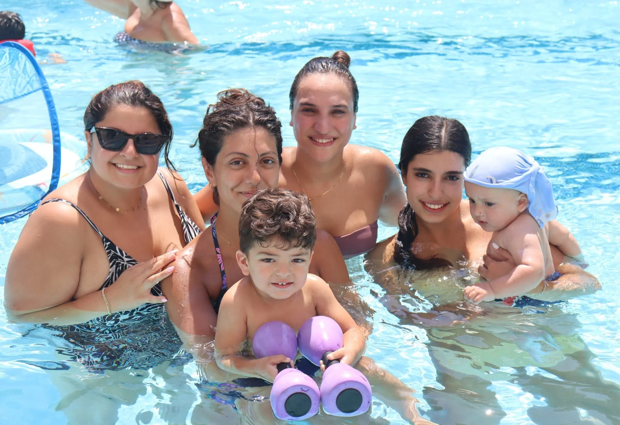 A group of people, including women and children, are in a swimming pool smiling at the camera on a sunny day, with some holding colorful balls.