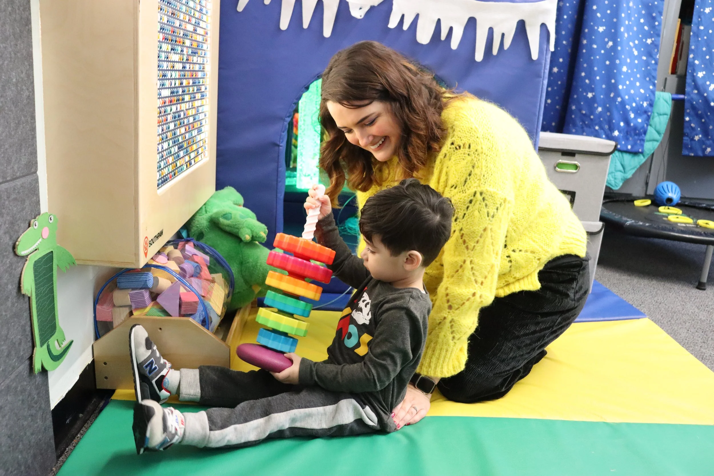 A woman with curly brown hair smiling and playing with a young boy in a colorful indoor play area. They are sitting on a green and yellow mat, engaging with a stacking toy. The background includes a blue wall decorated with white snowflakes and icicles, and various toys and play equipment.