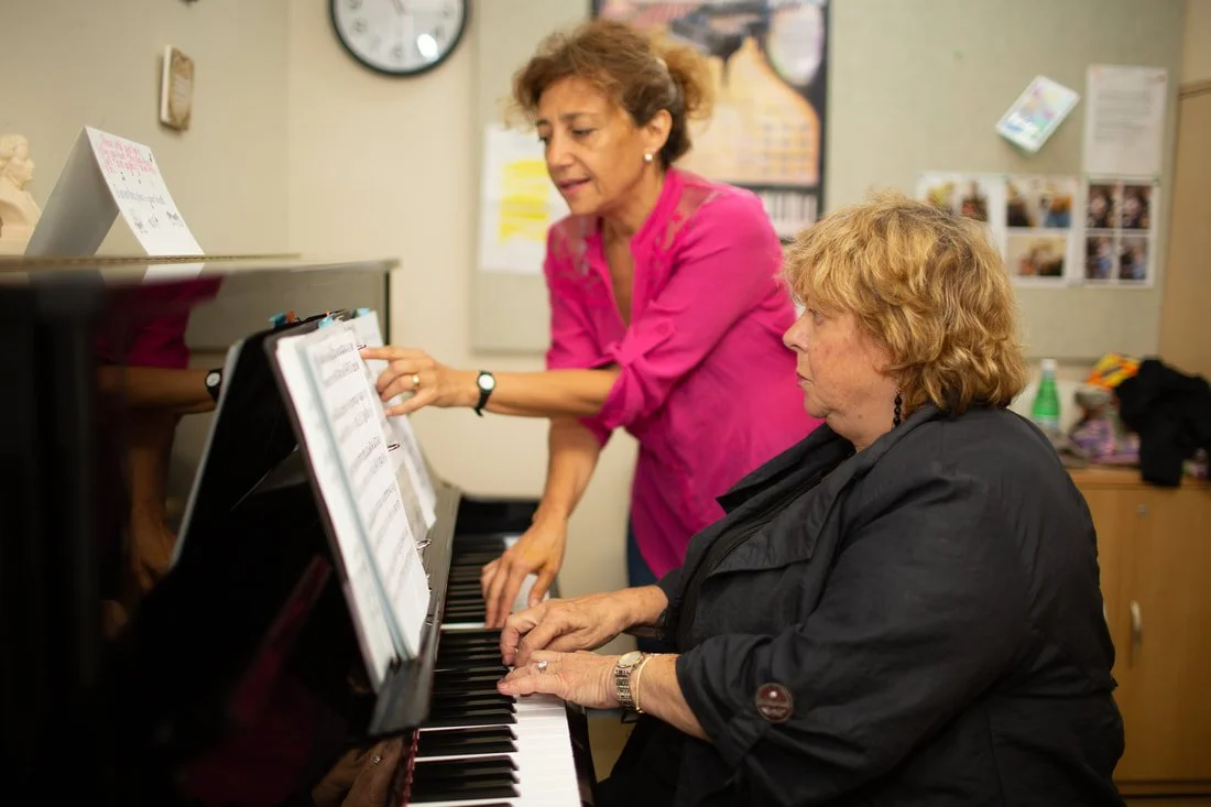 Two women are engaged in a piano lesson; one is playing piano, and the other is instructing her, in a classroom setting.