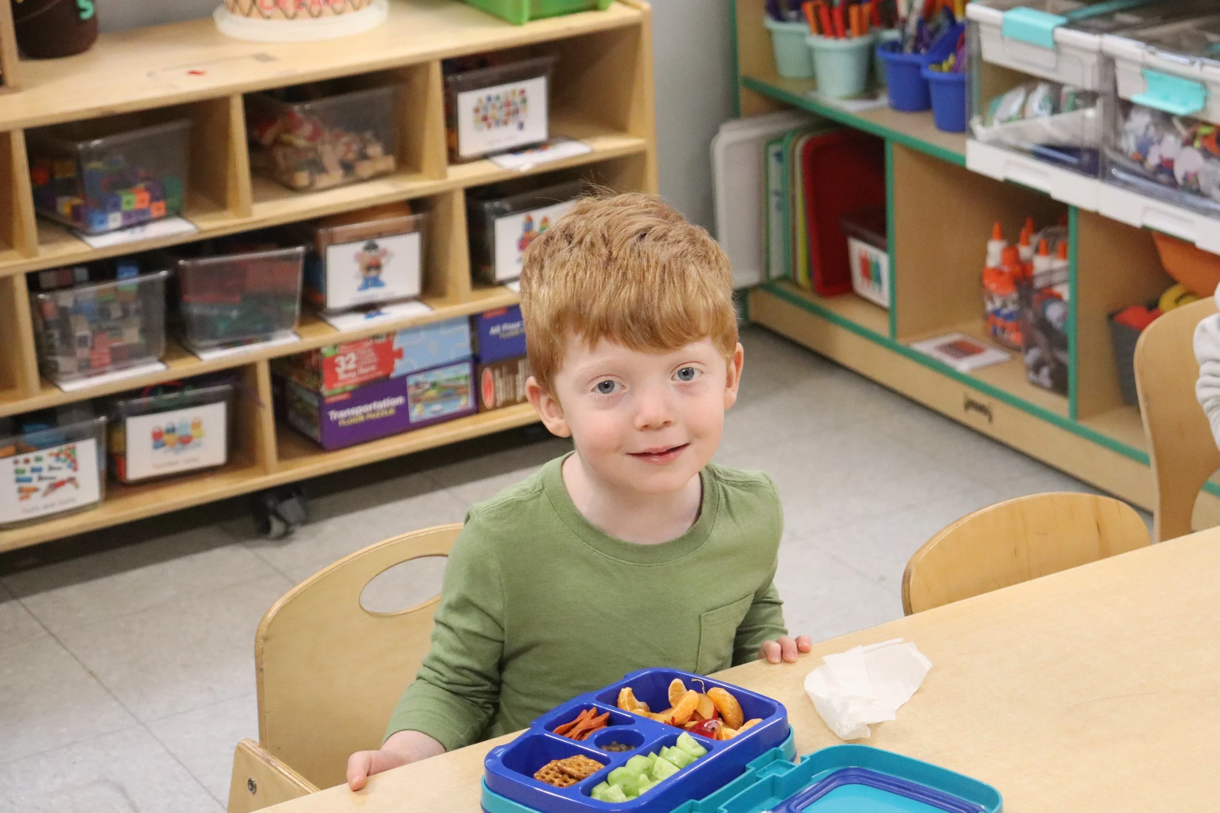 A young boy with red hair and blue eyes sitting at a table in a classroom, smiling at the camera. There is a blue divided tray with food items in front of him, including crackers, celery, and grape tomatoes, and a napkin next to it. Shelves with toys and educational materials are in the background.