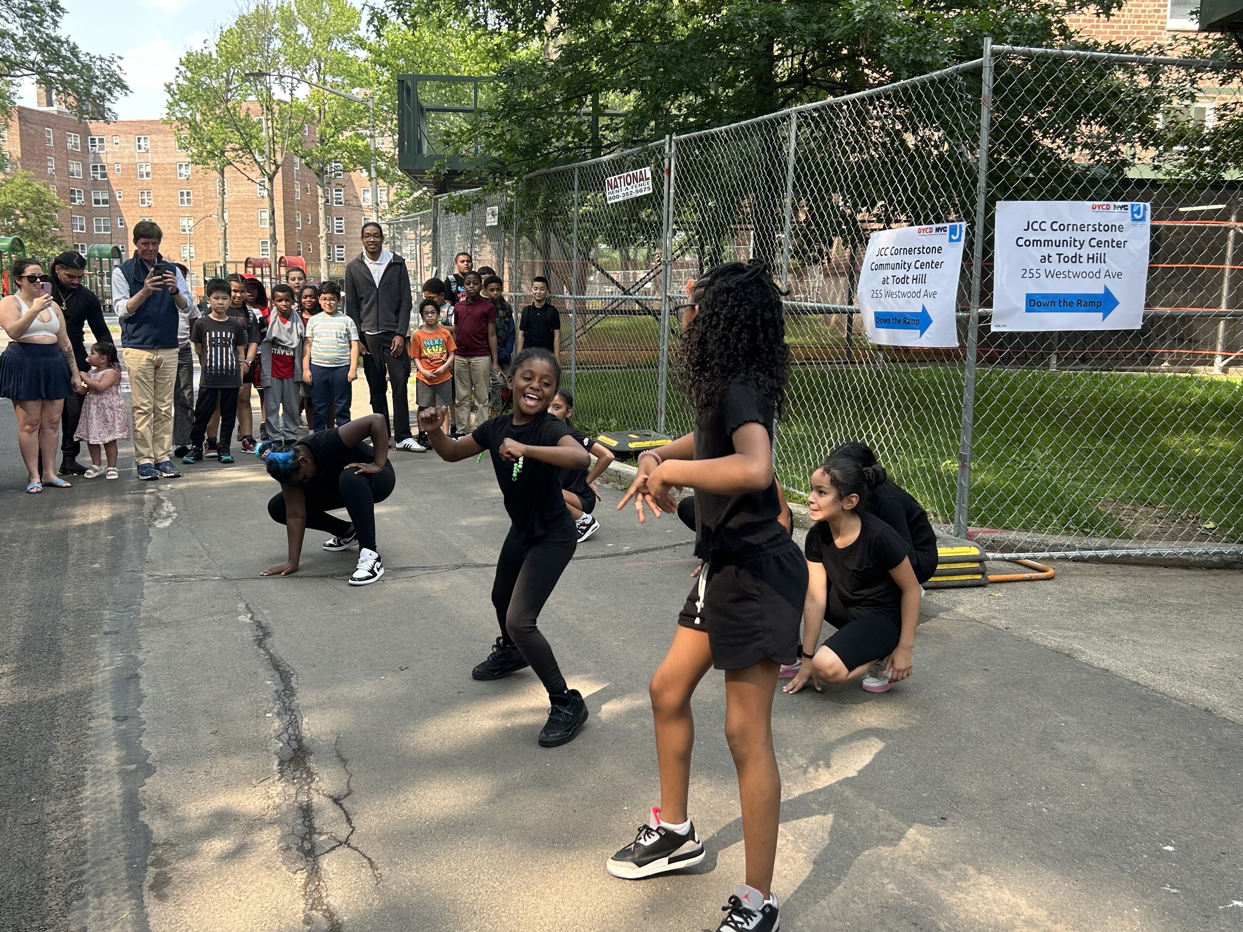 Children and adults at an outdoor community event in a park, with a chain-link fence, signs directing to JCC Cornerstone Community Center, and trees and apartment buildings in the background.