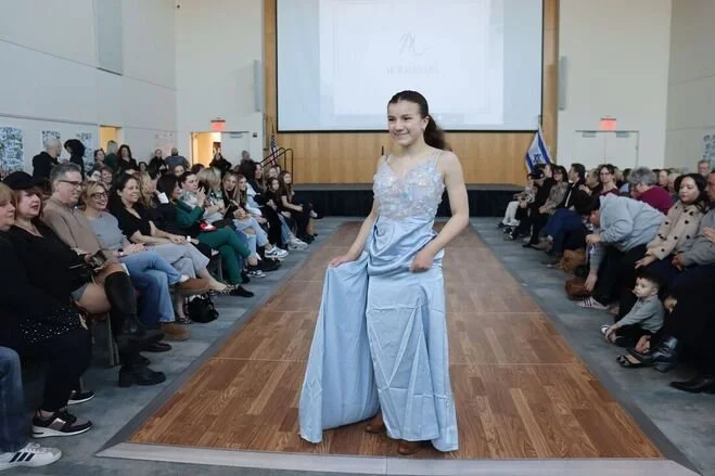 A young girl walking on a wooden runway at an indoor event, wearing a light blue dress. Audience members are seated on both sides of the runway, watching her. A large screen is visible in the background.