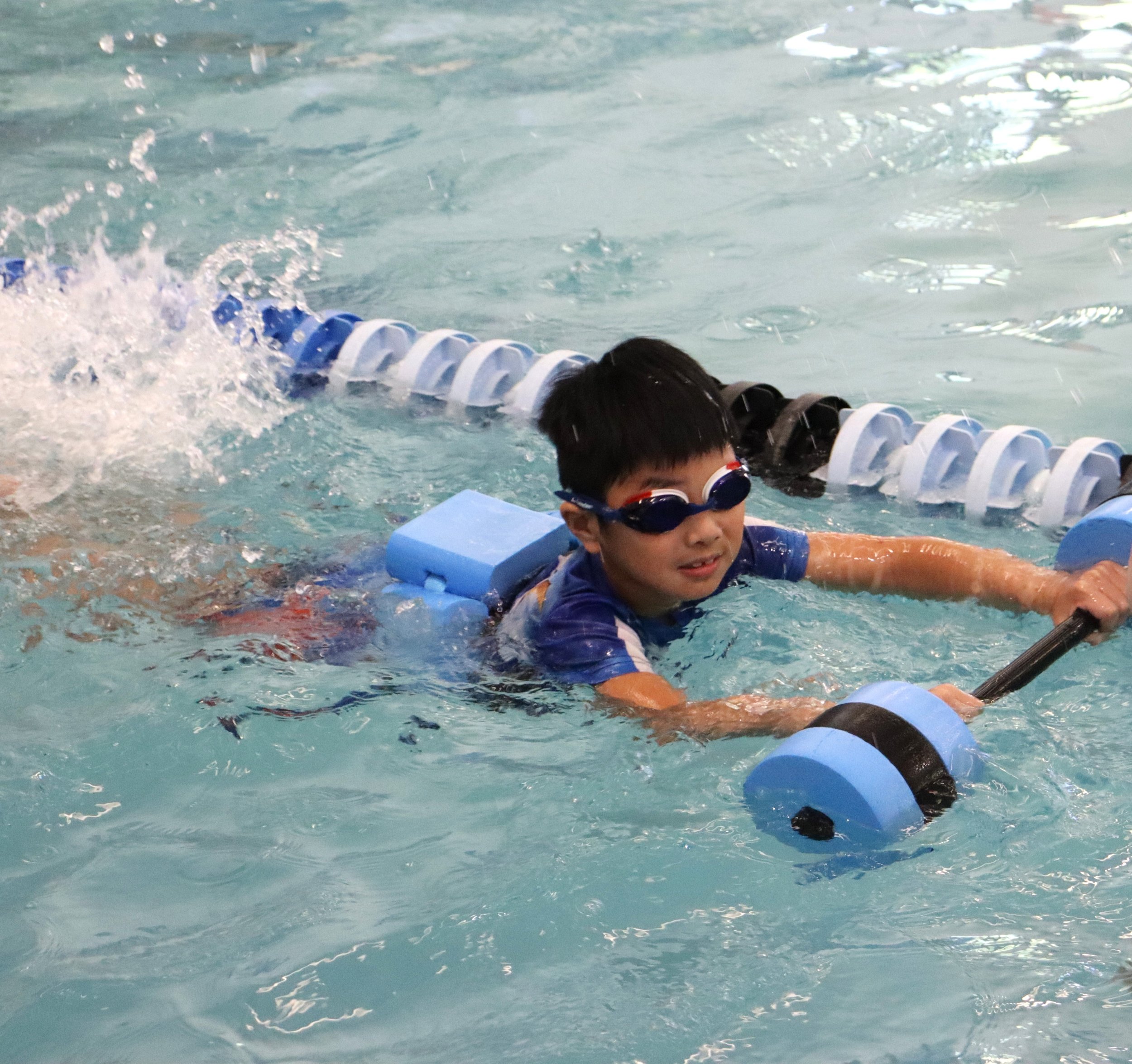 A boy swimming in a pool with goggles, holding onto a kickboard, and a lane marker in the water.