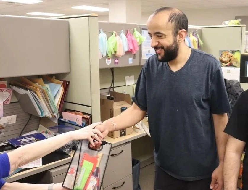 A man with a beard and dark hair, wearing a black T-shirt, shaking hands with a person in an office or store environment.