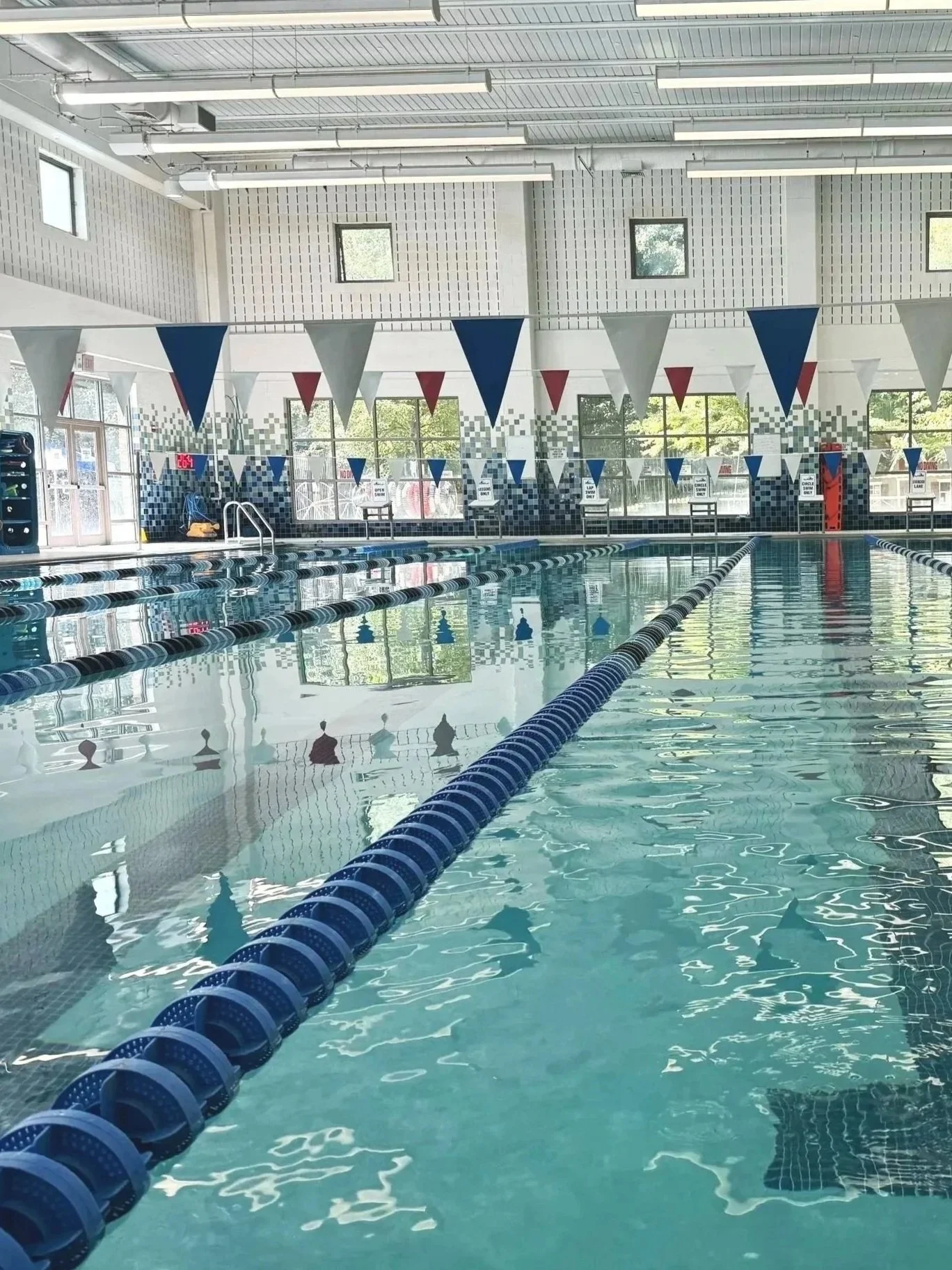 Indoor swimming pool with blue and white lane dividers, flags hanging from the ceiling, and windows letting in natural light.