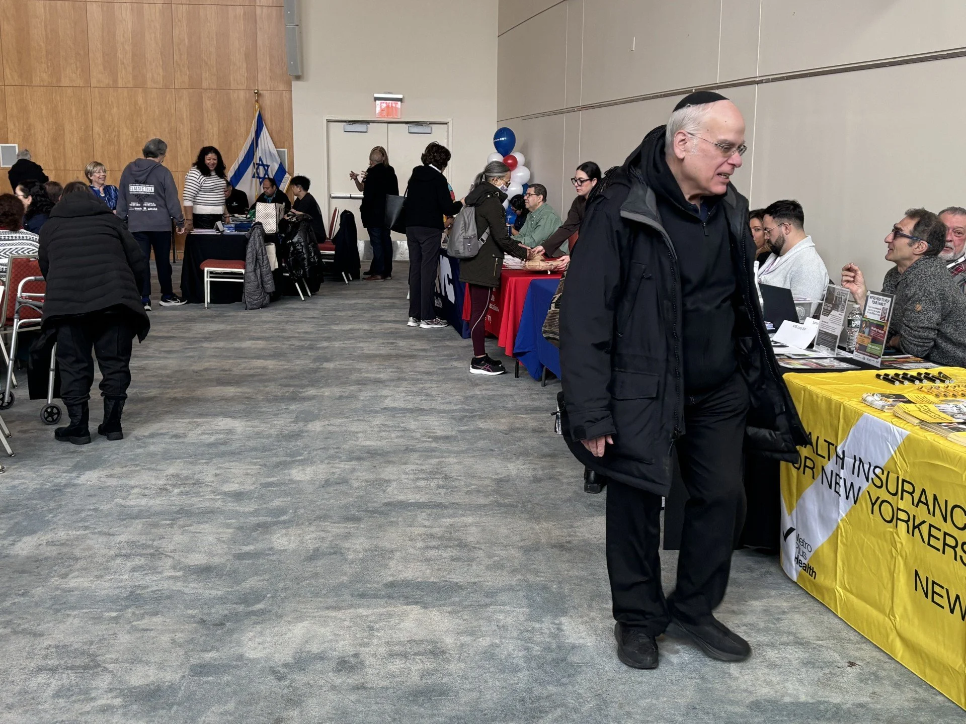 People attending an informational event or conference in a large indoor space with tables, display boards, and balloons. Some individuals are engaged in conversations and exchanging documents.