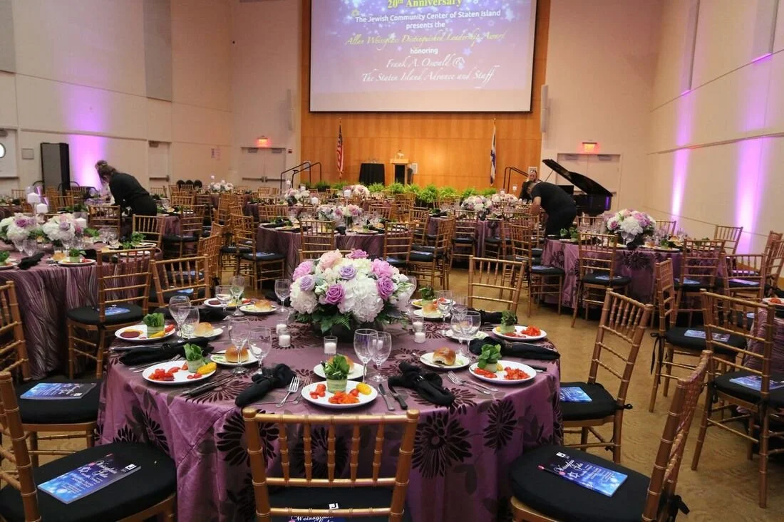 Banquet hall with round tables decorated with purple tablecloths and floral centerpieces, set for a formal event. Plates with food, glasses, and black napkins are arranged on each table. Several empty chairs are around the tables, and some staff are preparing the setting. A stage at the front features a large screen displaying event information and is flanked by American and state flags.