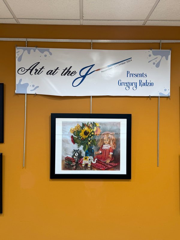 Art exhibit banner titled "Art at the J" presenting Gregory Radzio, featuring a framed artwork of a doll, a bouquet of sunflowers and other flowers, a small black and white animal figurine, and a decorative item resembling a robot or toy, all on a yellow wall.