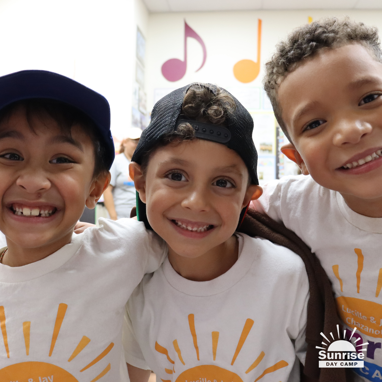 Three smiling children wearing white T-shirts with a sun logo and the text 'Sunrise Day Camp'. They are close together in a classroom with musical notes on the wall in the background.