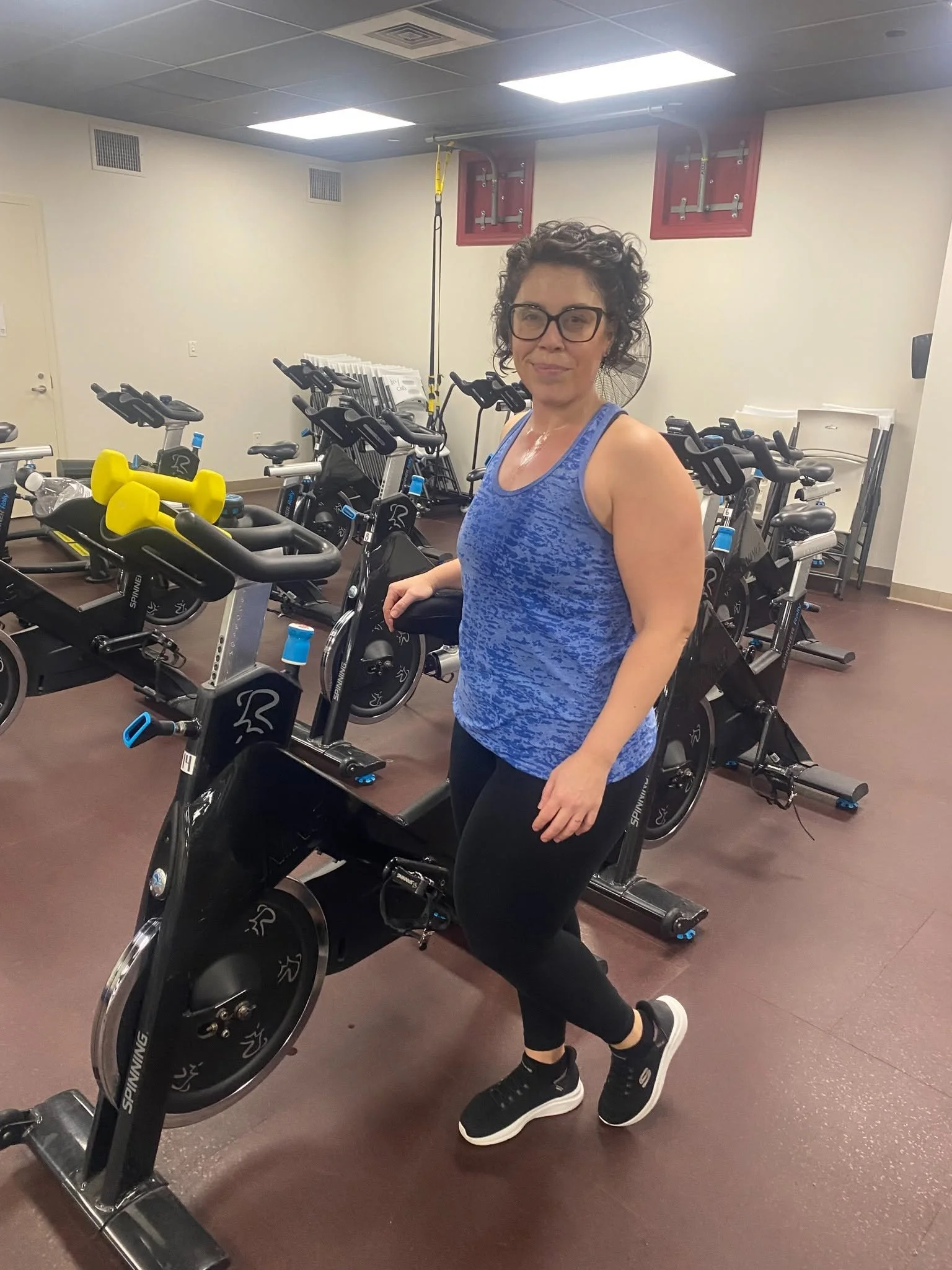 A woman with curly hair and glasses wearing a blue workout top and black leggings appears to be exercising on a stationary bike in a gym. The gym has multiple exercise bikes, a fan, and a ceiling with bright lights.