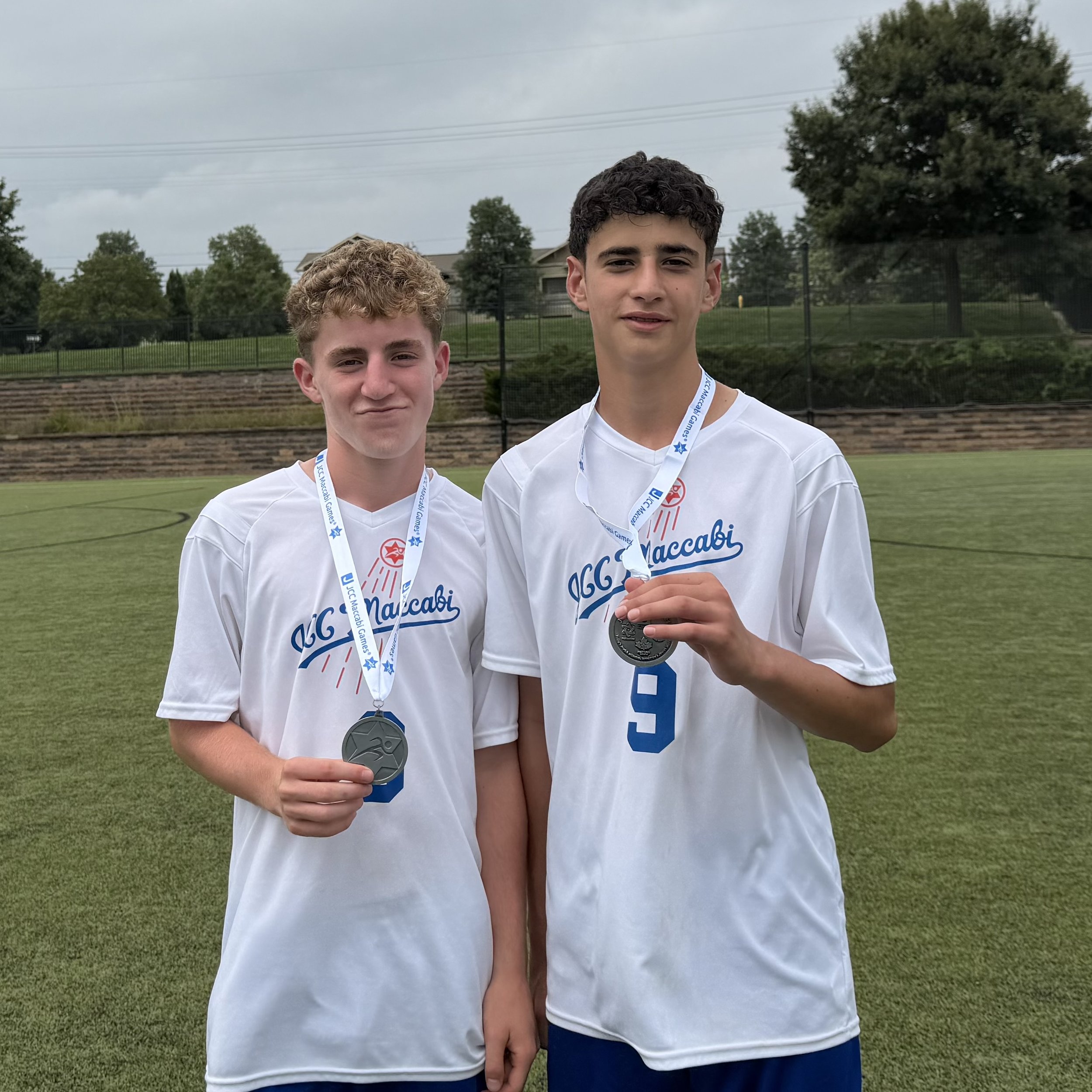 Two young men standing on a sports field, wearing white T-shirts with red and blue logos, each with a medal around their neck. One of the young men is holding his medal with his hand. They are posing for a photo on a cloudy day, with trees and residential houses in the background.