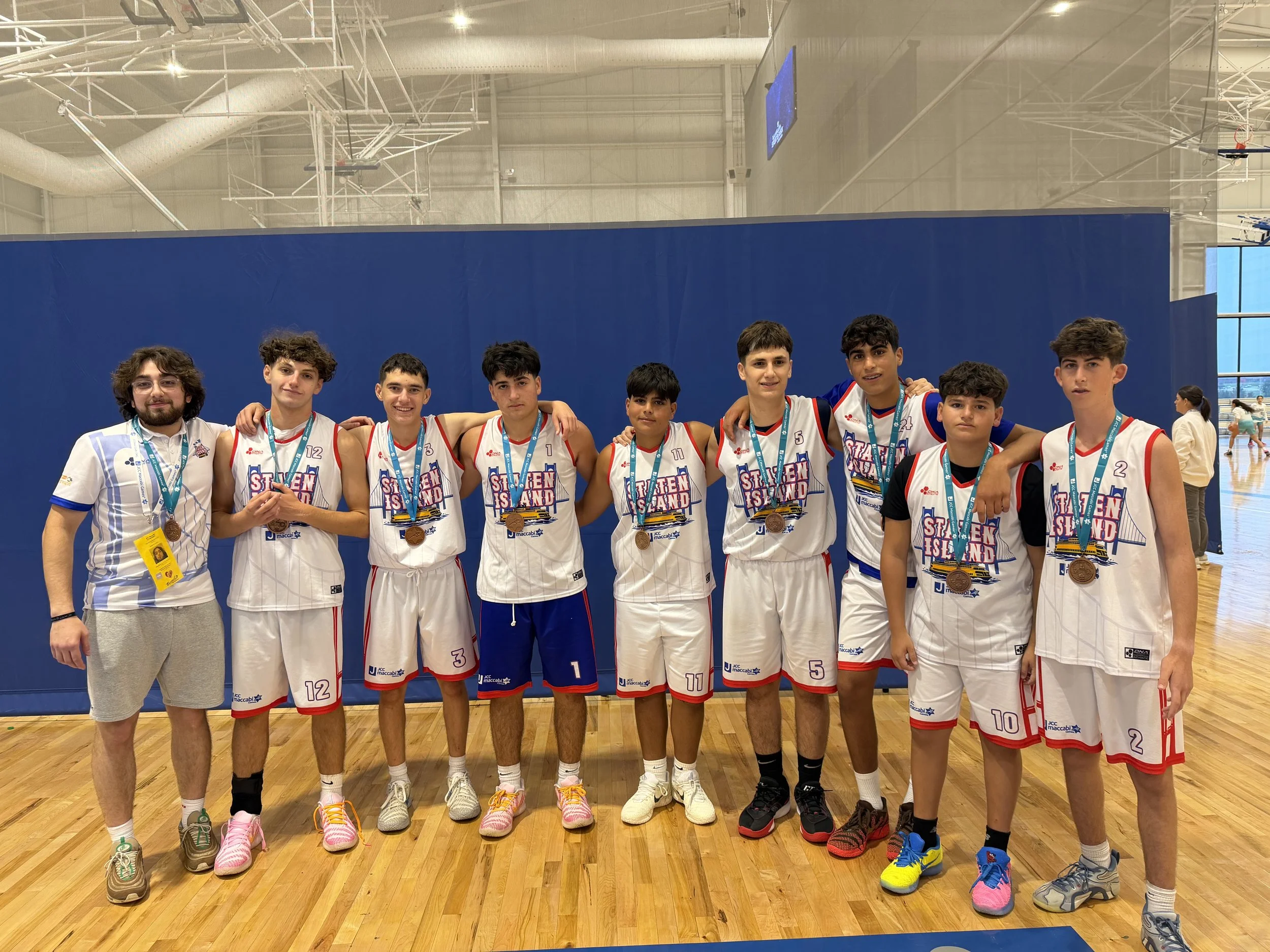 A group of young basketball players and one coach or team staff member standing together on a basketball court. The players are wearing white team uniforms with red and blue accents, medals around their necks, and sneakers of various colors. They are posing for a photo with their arms around each other, inside an indoor sports facility.