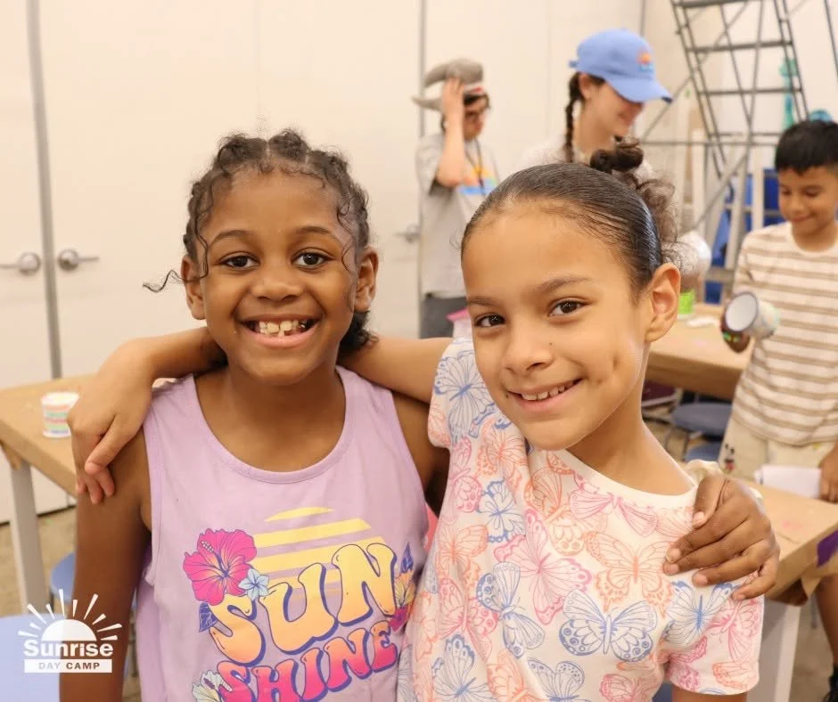 Two young girls smiling and hugging at summer day camp, with other children and counselors in the background.