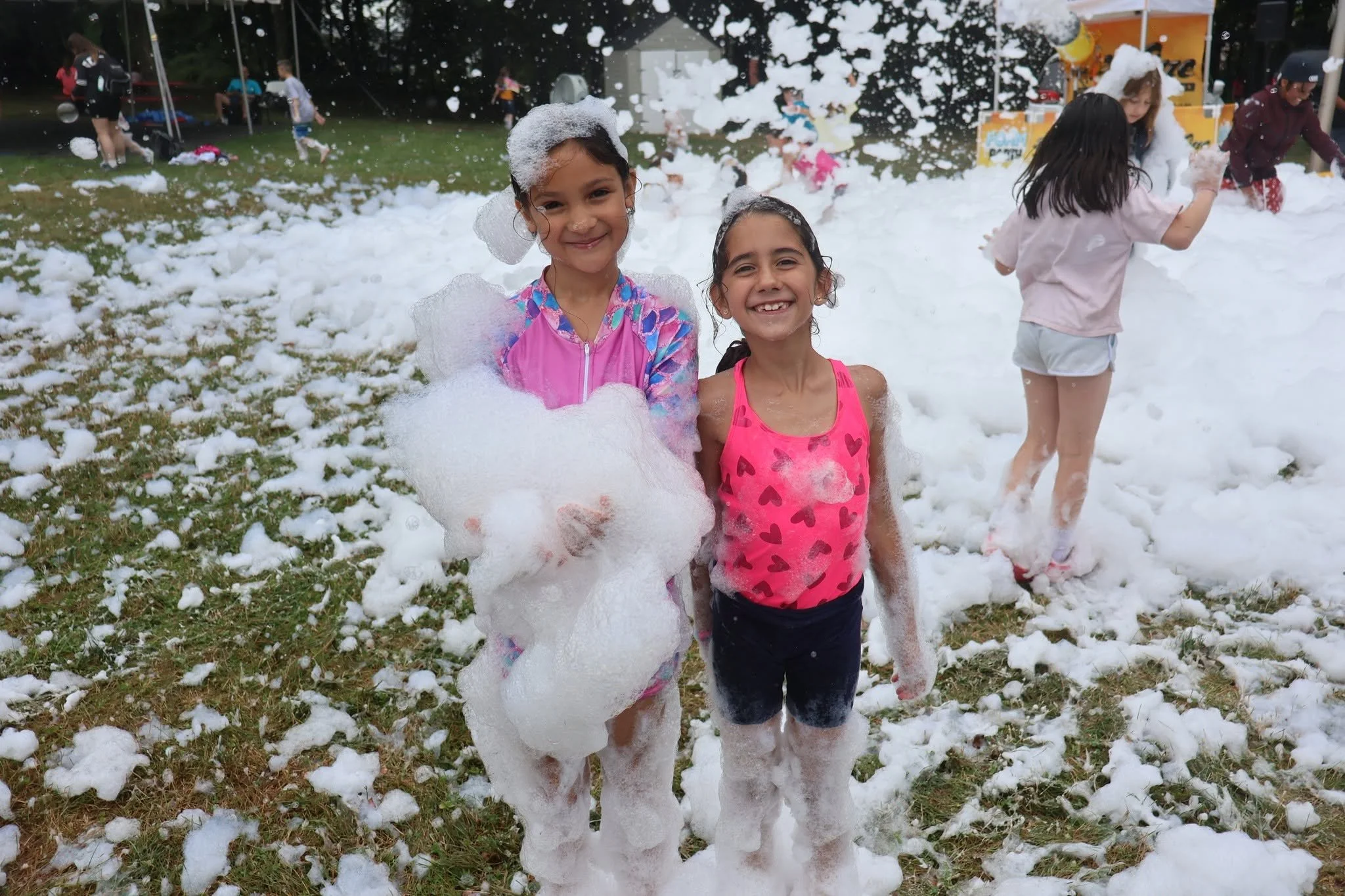 Two young girls smiling and covered in soap bubbles at a foam party outdoors, with other children and a yellow foam machine in the background.