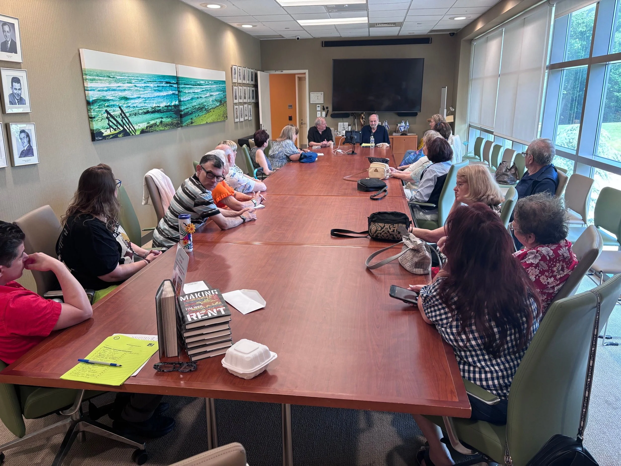 A group of people sitting around a large conference table in a meeting room. There are papers, books, and food containers on the table. The room has large windows on one side, letting in natural light. Four men are at the head of the table, with other attendees facing them.